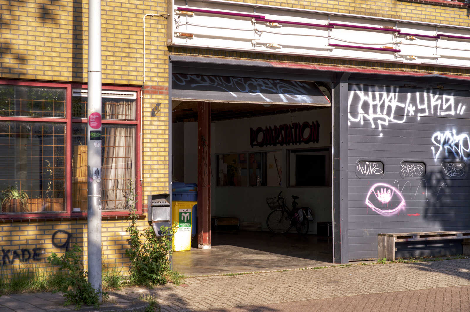 A gray building with graffiti and a metal awning. A bicycle is parked inside. A green exit sign and a waste bin are visible on the sidewalk. Lush greenery surrounds the area.