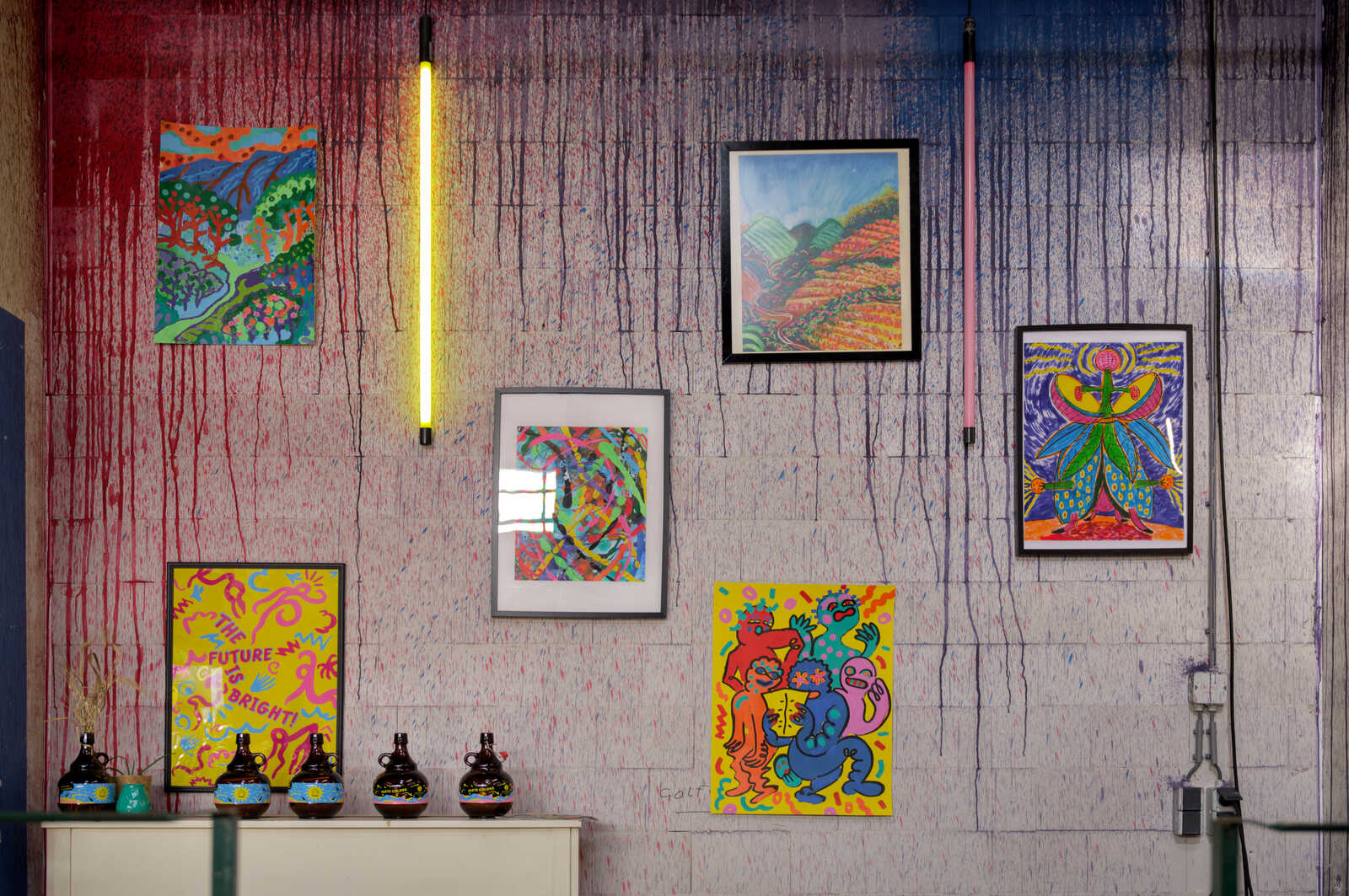A wall with colorful paint splatters showcases three framed, vibrant paintings and a yellow light fixture. Three dark bottles sit on a gray surface below the art. A sign reads The Future is Bright.