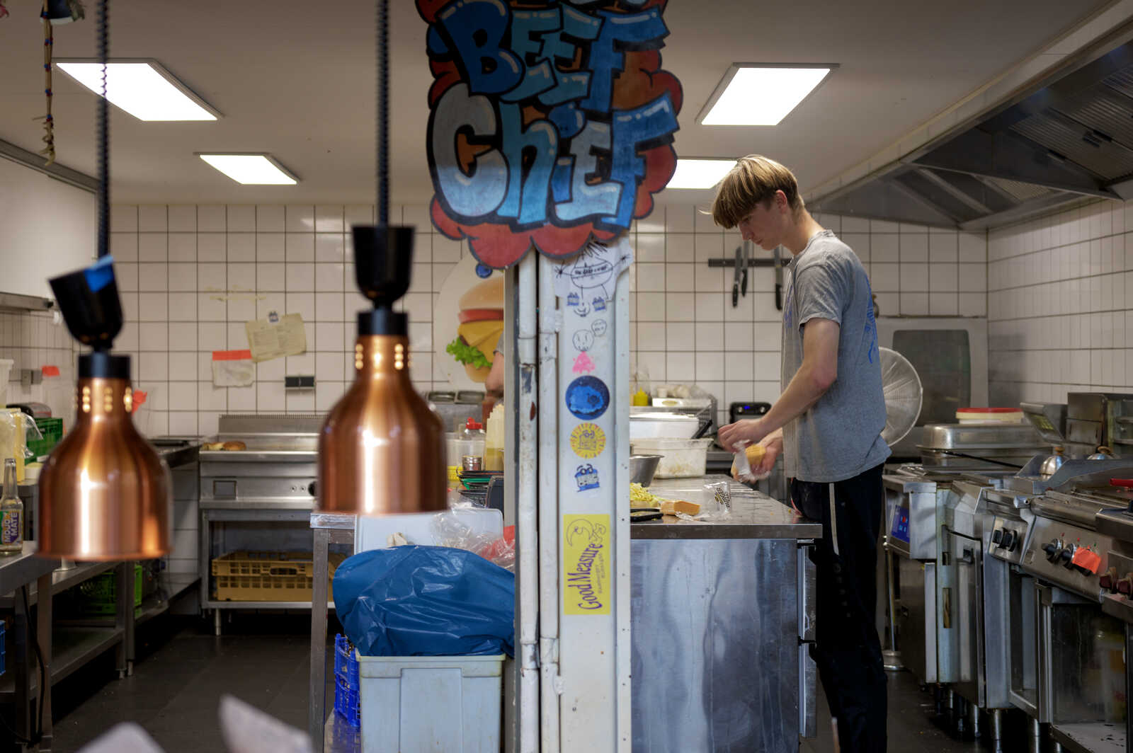 A person in a black shirt is preparing food at a stainless steel counter in an industrial-sized kitchen. Copper pendant lights hang above. Theres a fan and various kitchen utensils visible.