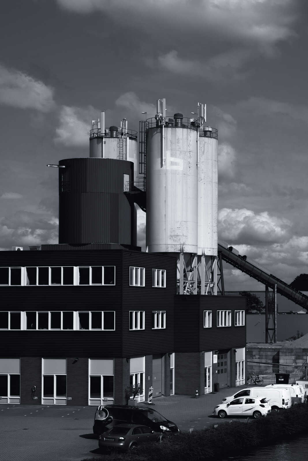 A tall, cylindrical silo with a ladder stands next to a dark, multi-story building. A loading conveyor leads to the silo, with parked cars and vans below against a cloudy sky.