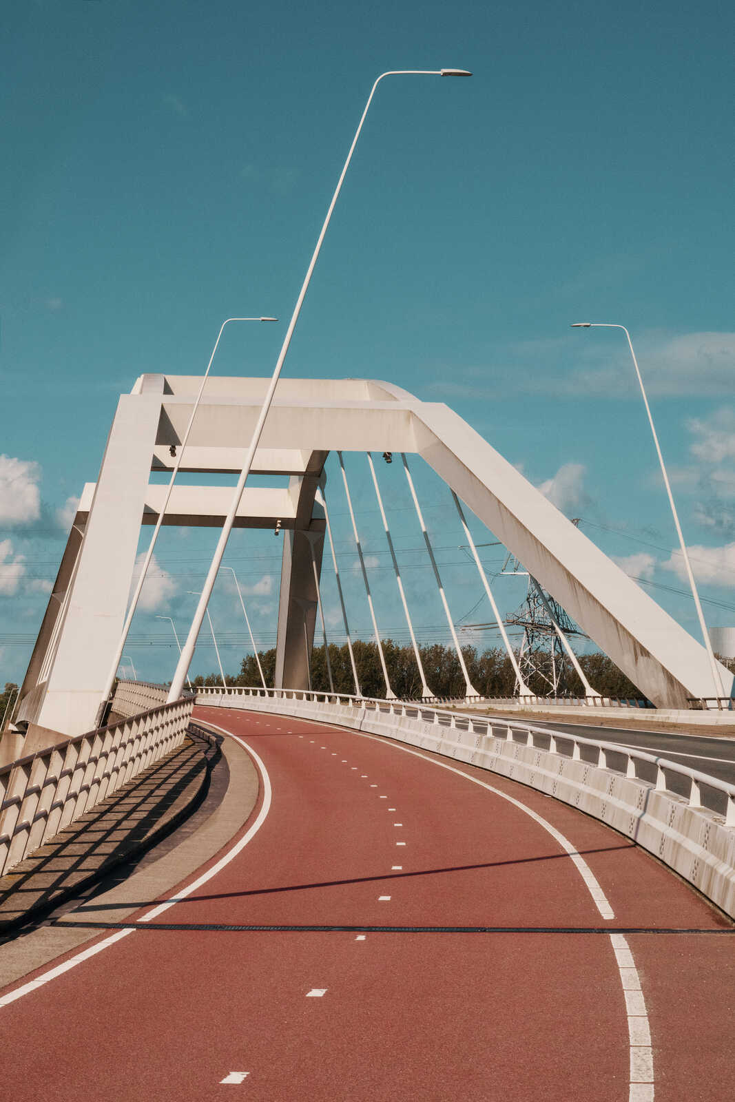 A long, arched white bridge spans a body of water under a blue sky with scattered clouds. White lines mark the roadway and pedestrian/bike path. Tall light poles line the bridge.