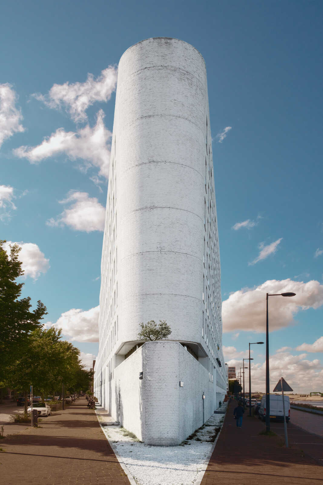 A tall, white, cylindrical building stands on a paved street with a sidewalk. Blue sky with scattered clouds. A tree grows near the buildings base. Cars are visible in the background. A pedestrian walks along the sidewalk.