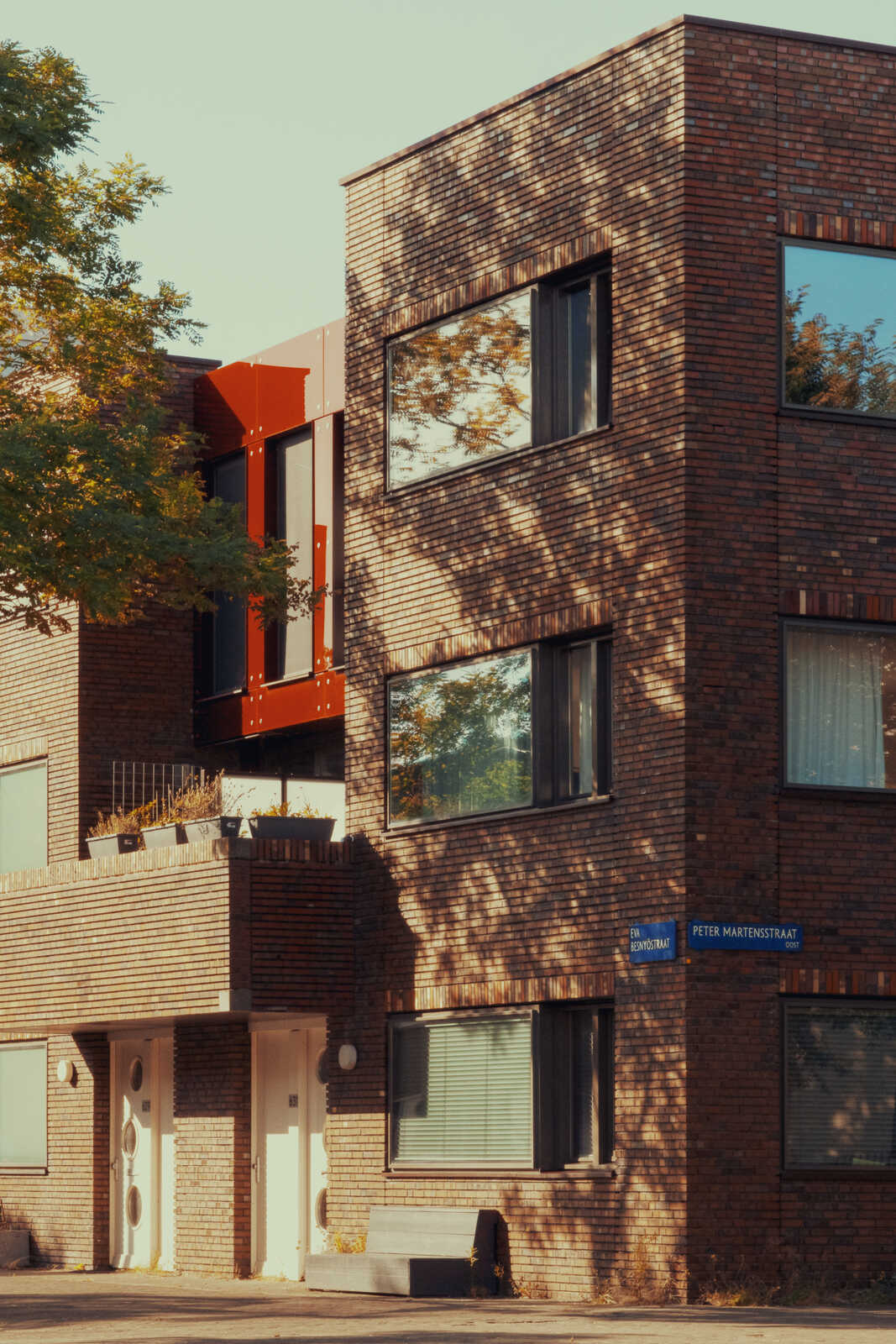 Red-brick building with dark-framed windows and a street sign reading Peter Martensstraat. A tree casts a shadow on the facade. Two white doors are visible at the base. Flat roof with a terrace.