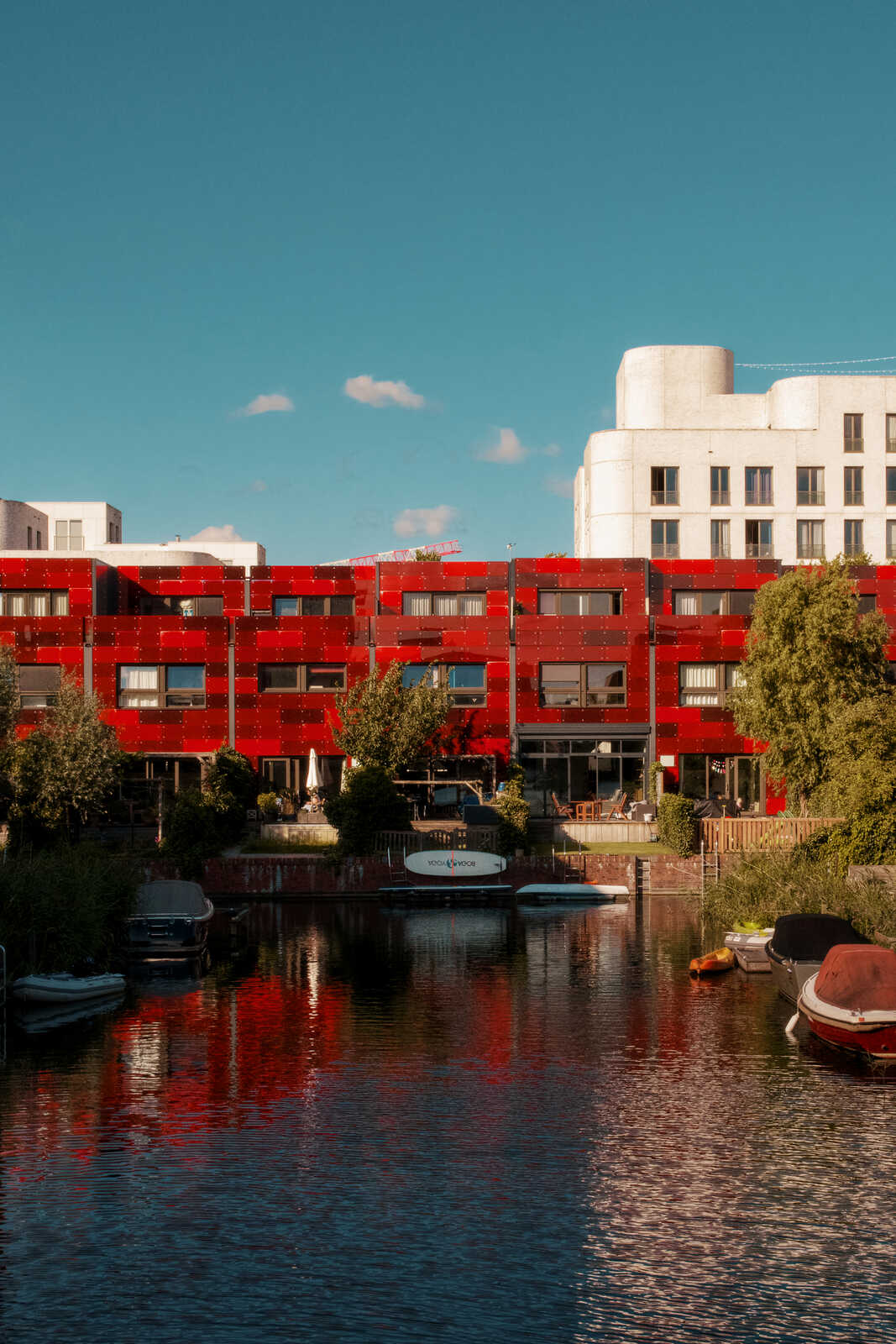 Red brick apartment building with dark green trees and a canal. Several boats are docked alongside the water, reflecting on the calm surface. A cloudy blue sky overhead.