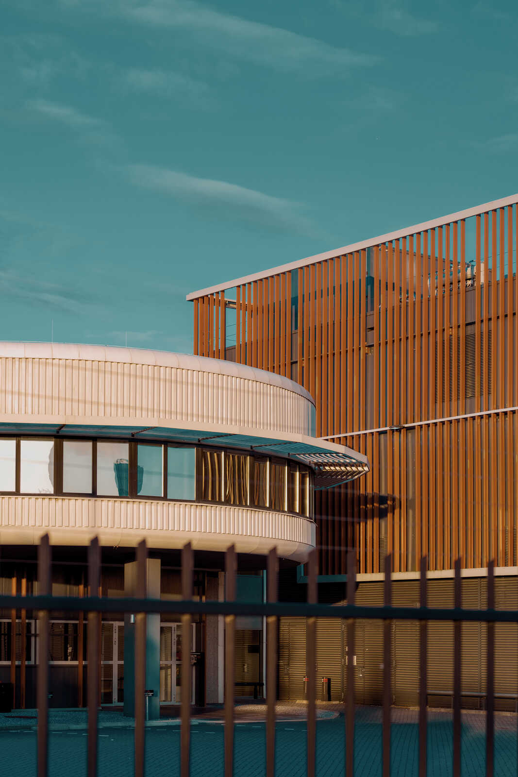 Modern building exterior with curved white facade and vertical wooden slats. Gray metal fence in foreground, blue sky backdrop, and visible windows. Street and sidewalk are also present.