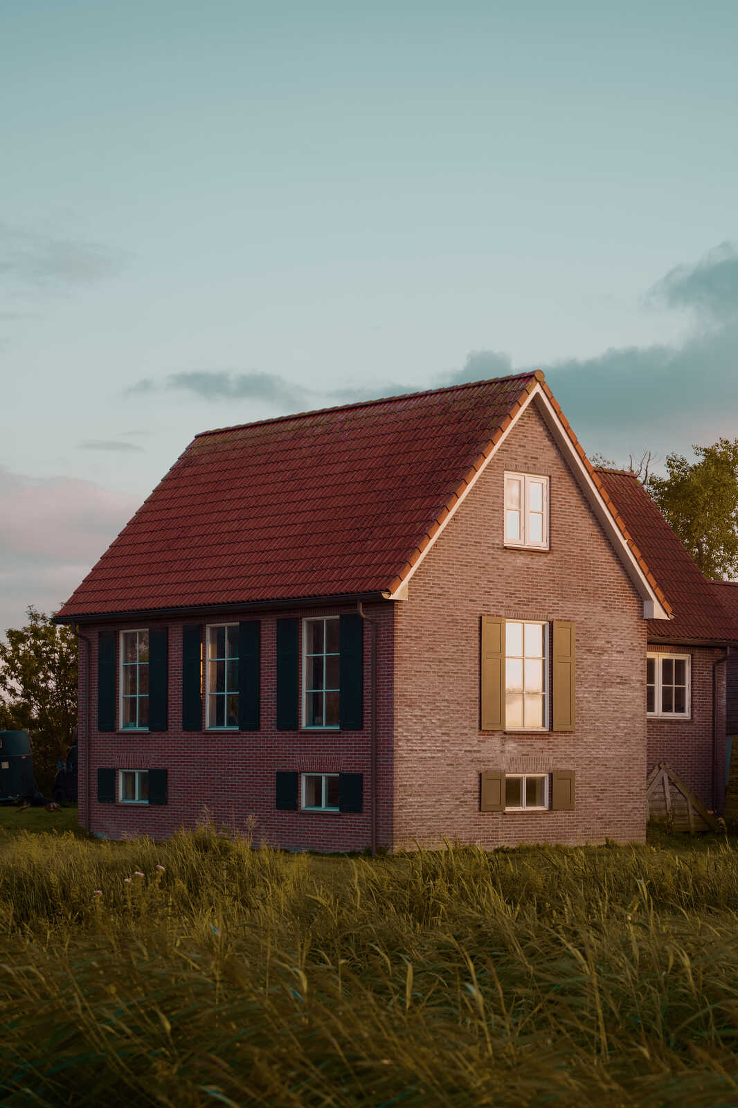 A two-story brick house with a red tile roof stands in a field of tall grass. Black-framed windows and light beige trim. The sky is a mix of pink and orange hues.