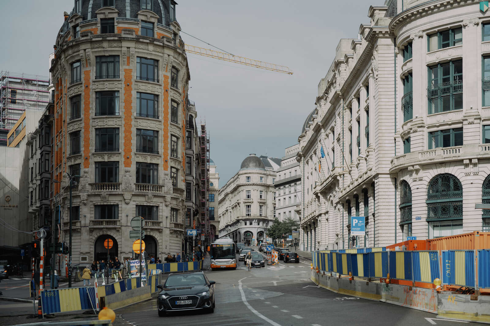 A street scene with ornate buildings, a yellow bus, a blue car, construction barriers, and a crane in the background. A pedestrian is visible on the sidewalk.