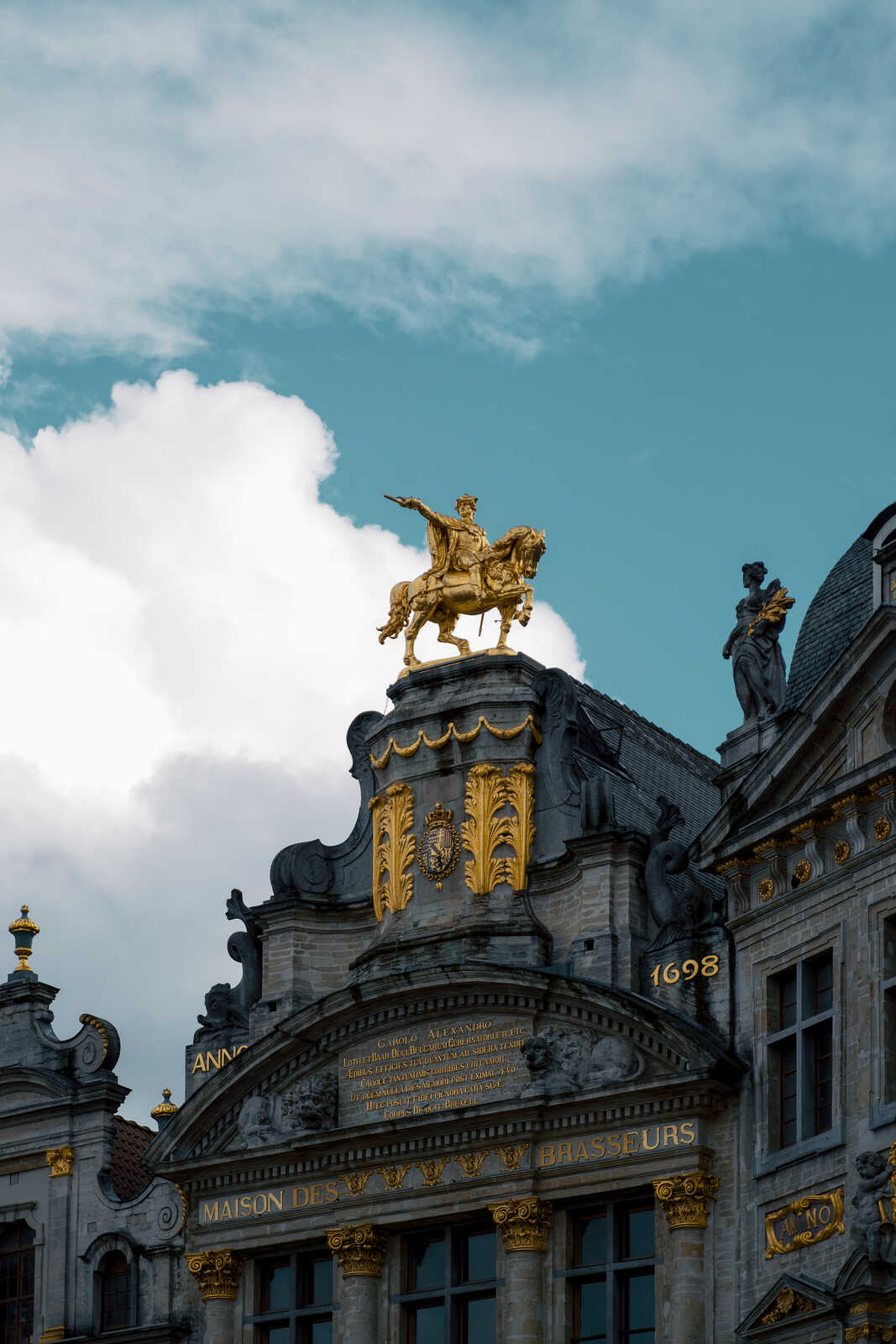 Ornate building facade with gold details and statues. The buildings name “Maison des brasseurs” is visible, along with the year 1688. A cloudy blue sky is in the background.