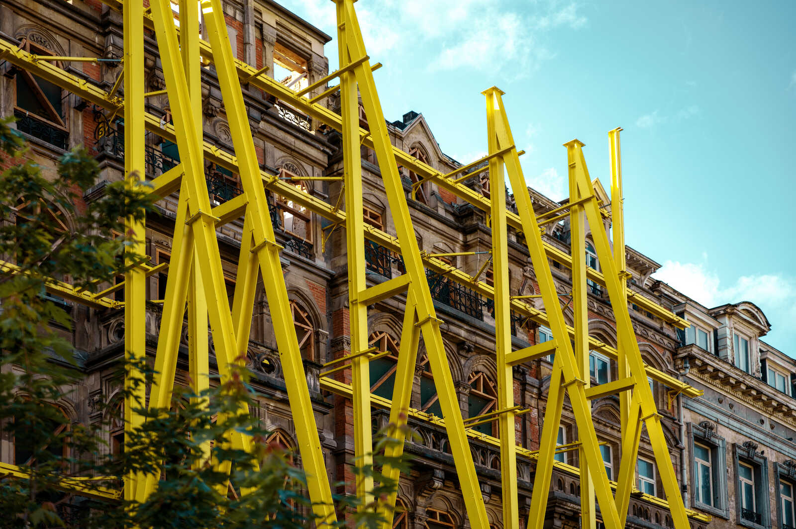 A tan stone building with arched windows is partially obscured by tall, bright yellow beams. A tree with green leaves is visible in the lower left corner, and a blue sky is above.