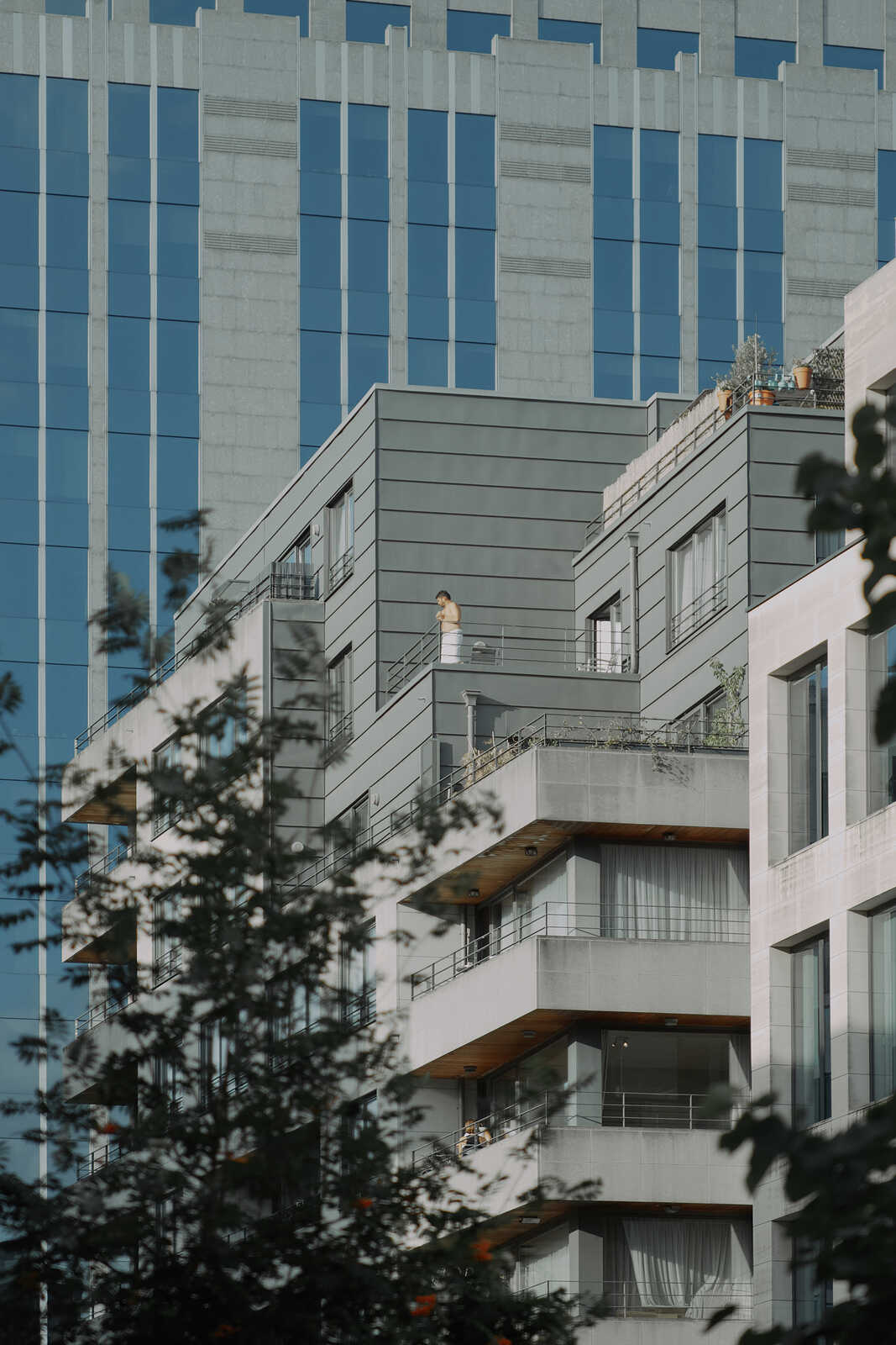 Gray building with multiple balconies and large windows. A figure stands on a balcony. Lush greenery is visible in the foreground. Blue sky in the background.