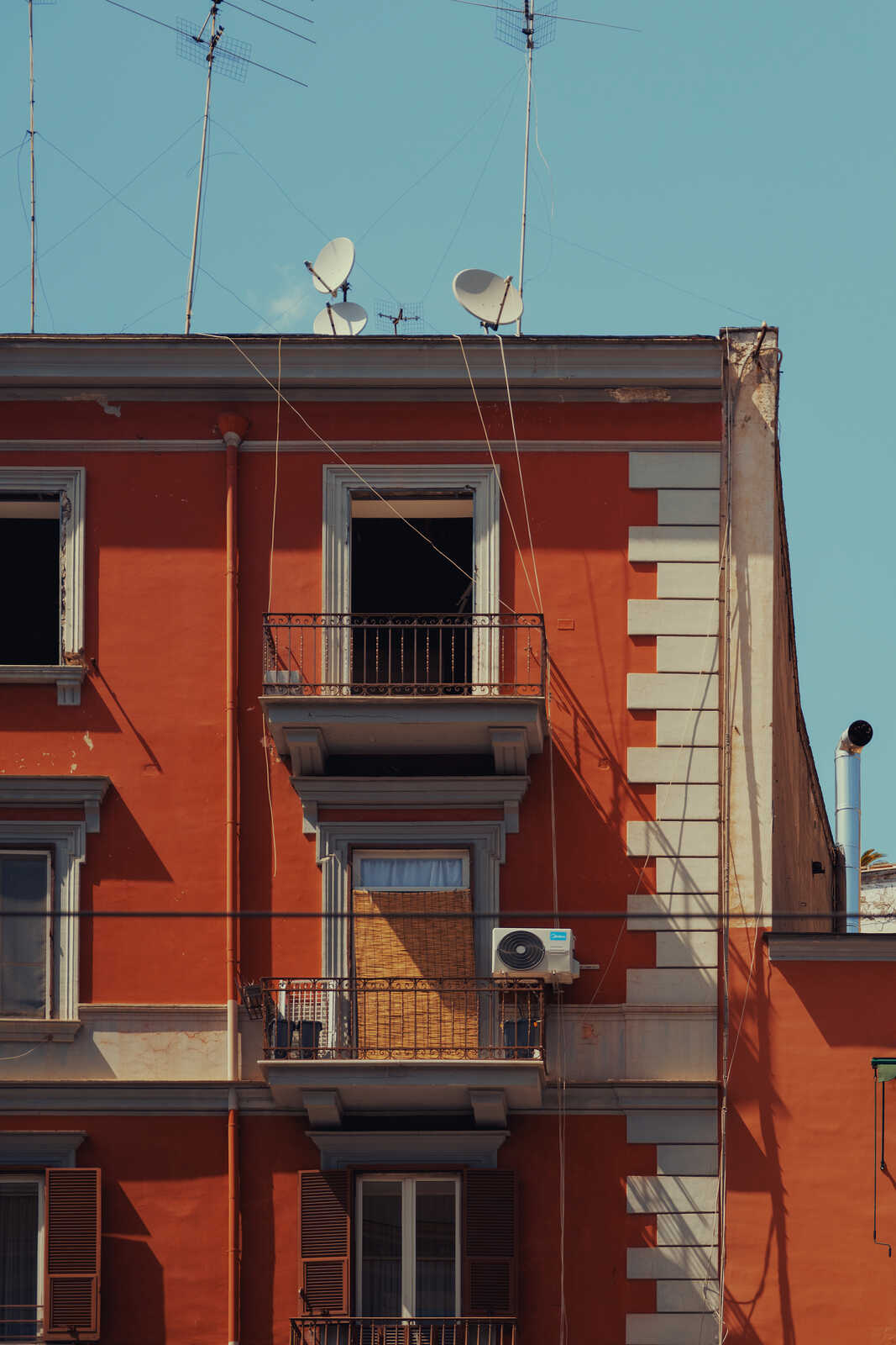 Red building with three windows and two balconies. Satellite dishes and antennas on the roof. A ventilation unit is mounted on the exterior wall. Blue sky in the background.