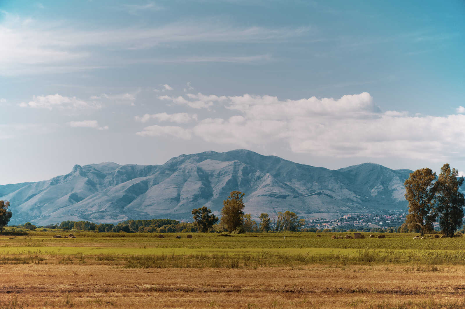 Rolling hills with green and yellow fields, scattered hay bales. Distant gray mountains rise under a partly cloudy blue sky. Trees line the horizon and a town is visible.