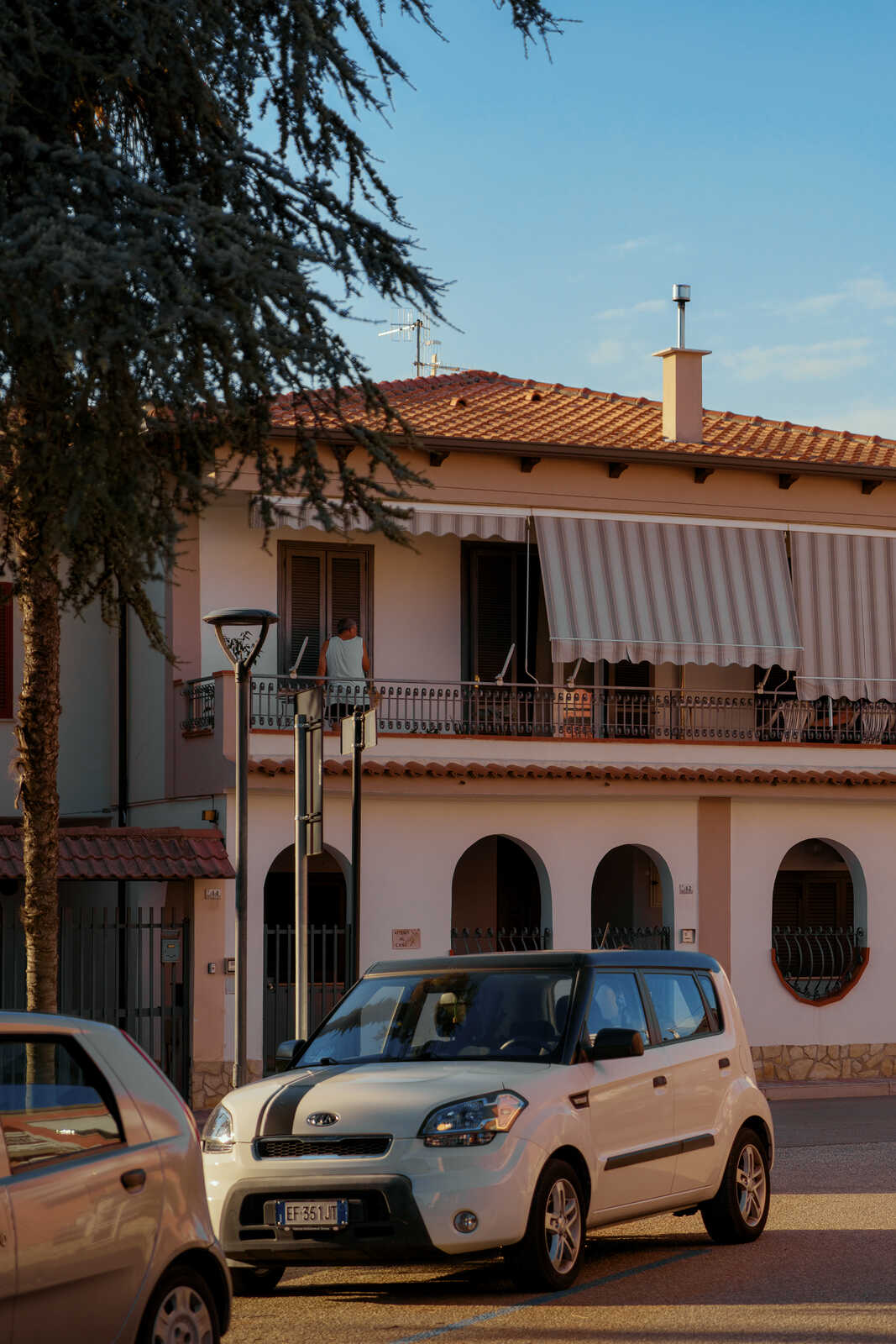 A white Kia Soul sits on a street. A building with arched windows and a balcony is behind it. A tree stands to the left, and a blue sky is overhead.