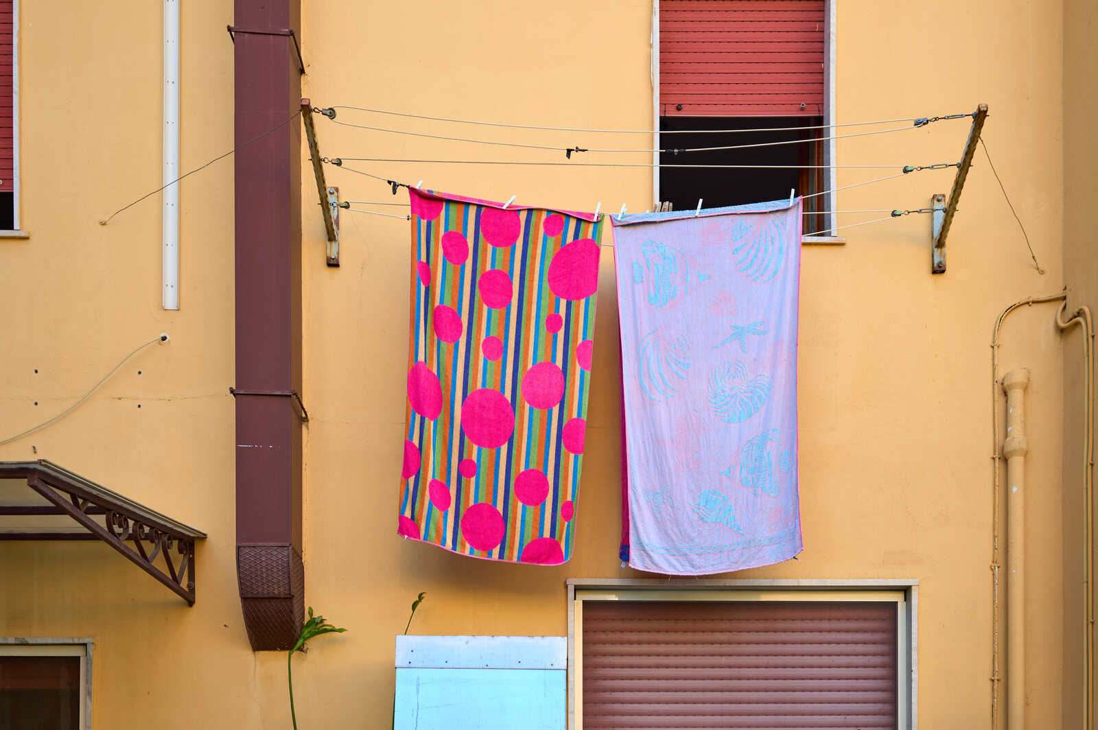 Two towels hang on a clothesline against a yellow wall. One towel is pink with dark blue and green dots. The other is light blue with a floral pattern.