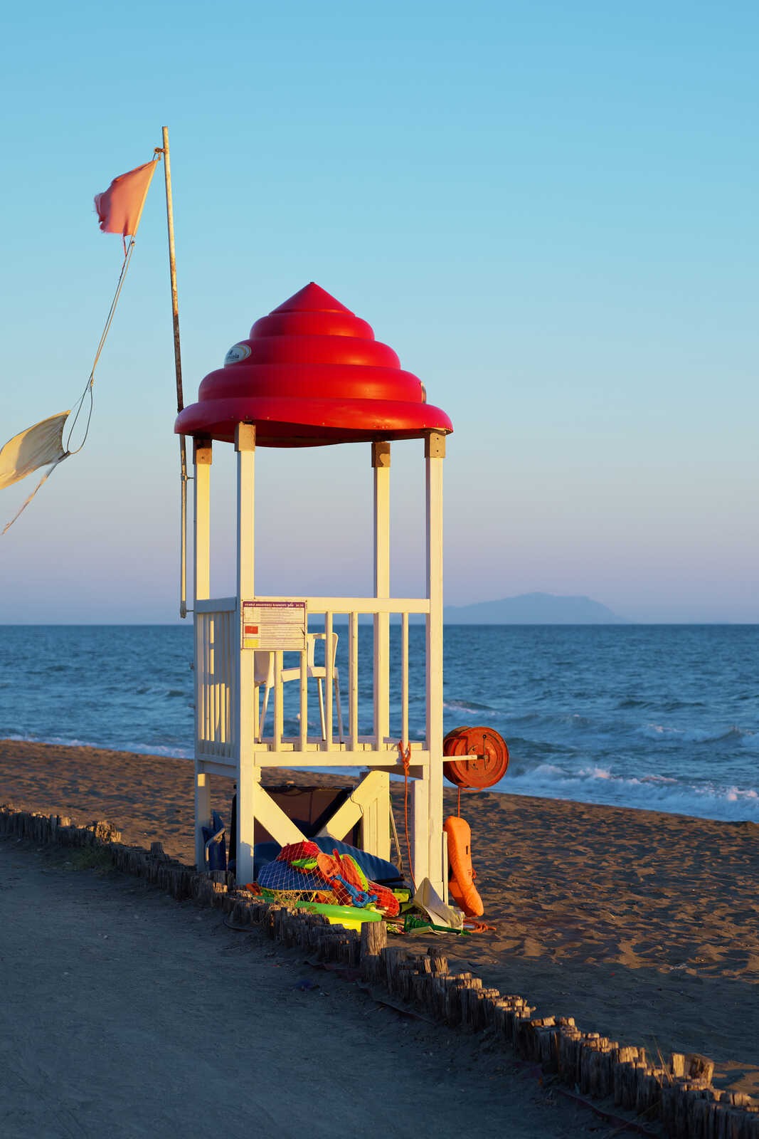 A white lifeguard stand sits on a sandy beach. A red rescue buoy is mounted to the side. The ocean and a distant shoreline are visible in the background. A flag waves to the left.