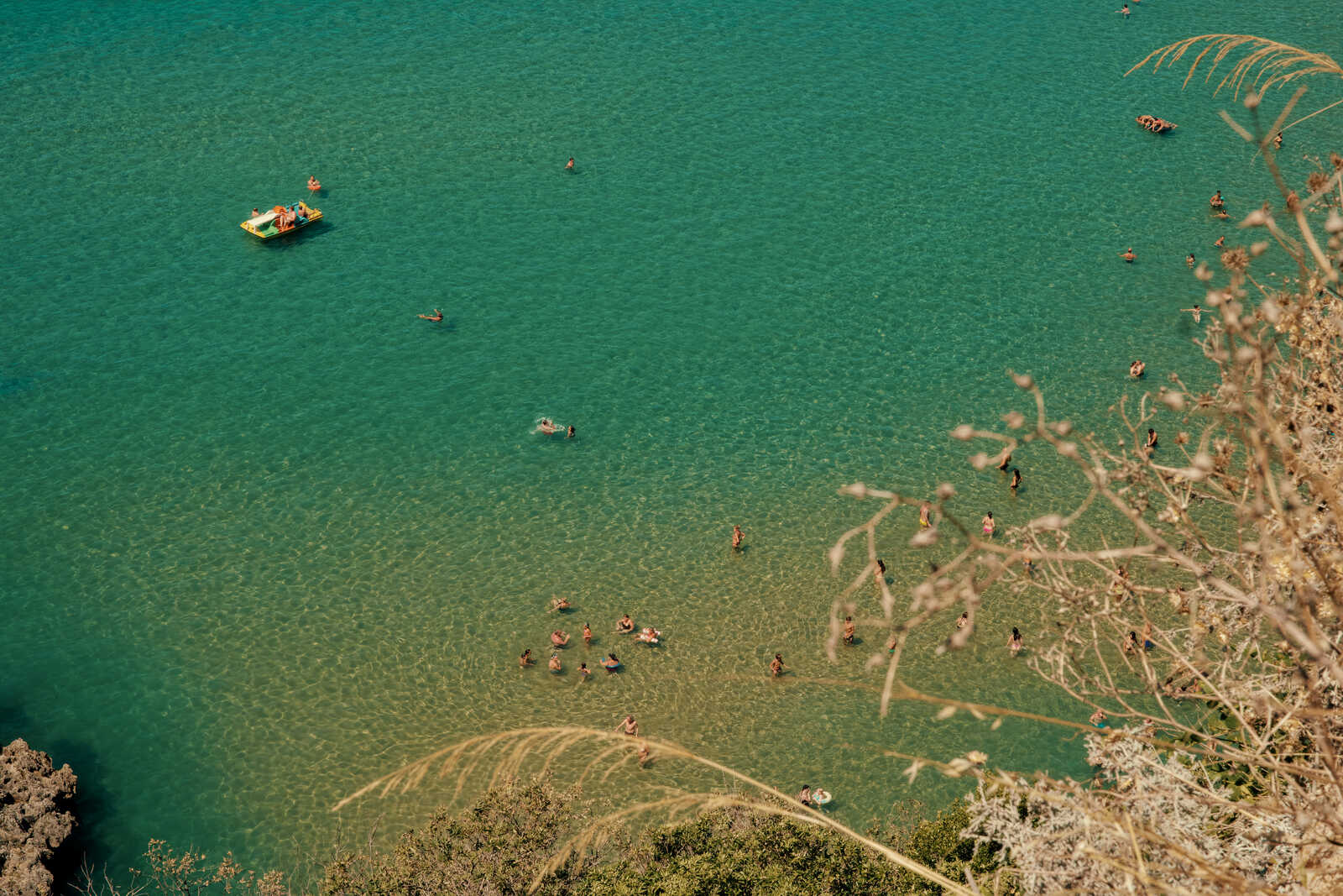 Aerial view of turquoise water with numerous figures swimming and floating. Some figures are on a small inflatable raft. Dry grass and branches are visible in the foreground.