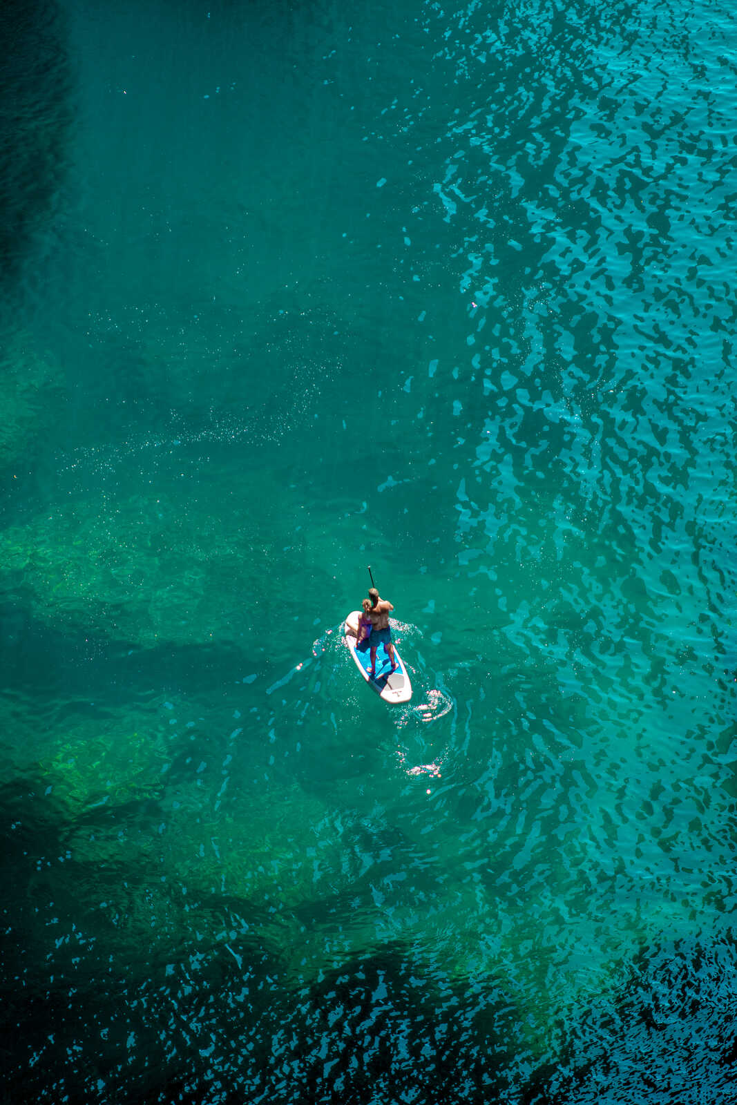 A person paddles a white stand-up paddleboard on turquoise water. Ripples stretch out from beneath the board. The water has a textured, shimmering surface.