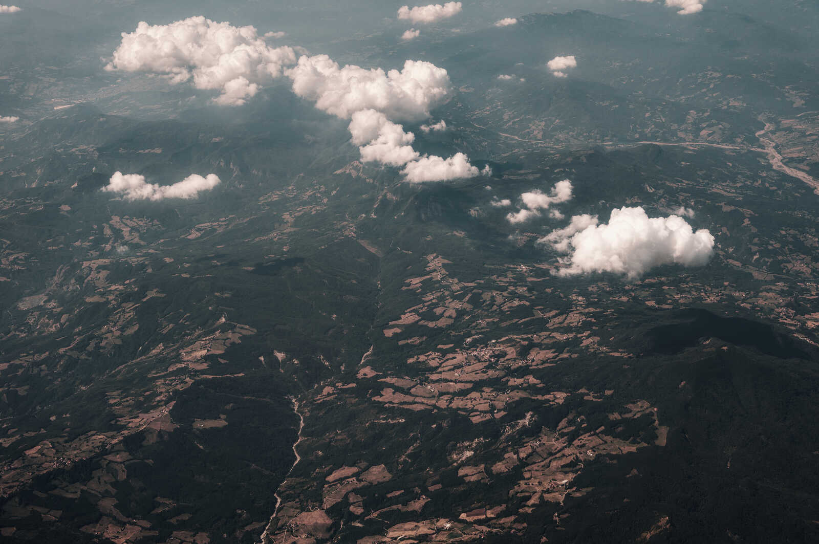 Aerial view of a dark, forested mountain range. Scattered white clouds float above. A few roads and small structures are visible within the landscape. Grayish-blue sky.