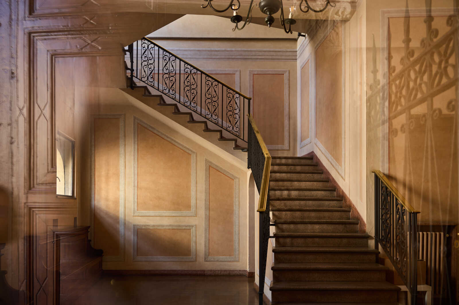 A grand staircase with ornate iron railings ascends. Beige-toned walls feature paneling, and a chandelier hangs above. A mirror and a table are visible on the left. The stairs are carpeted and have a wooden banister.