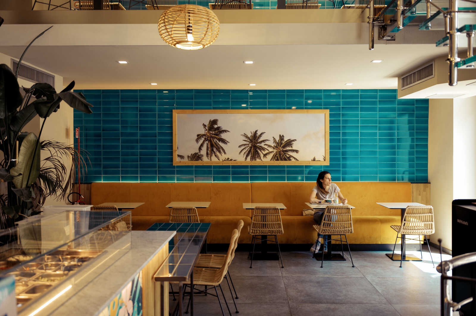 A cafe interior with turquoise tiled walls and rattan furniture. A person sits at a table, a palm tree print hangs above a yellow bench. A wicker pendant light is visible. A refrigerated display case is in the foreground.