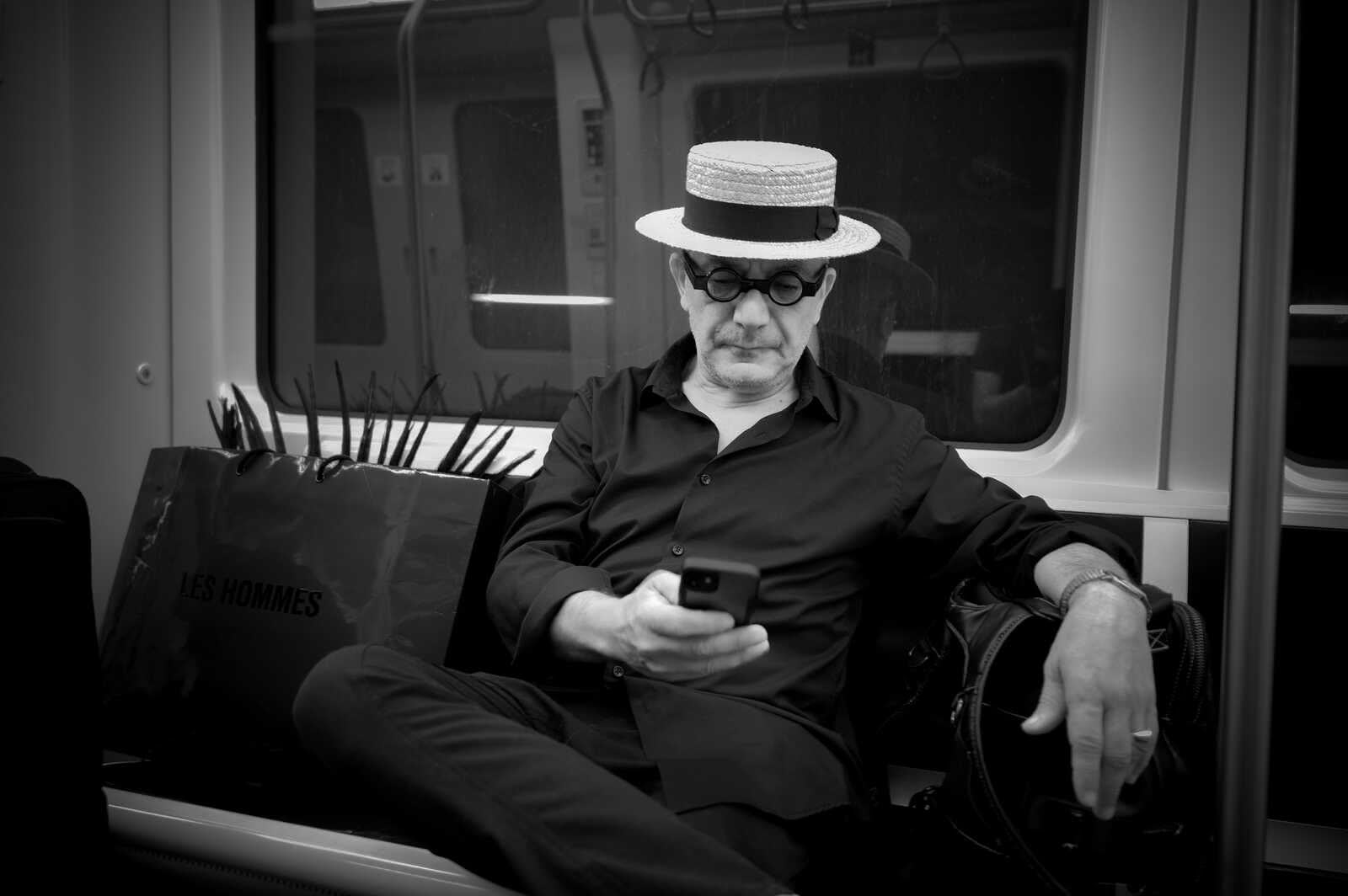 Black and white photo of a man wearing a straw hat and glasses sitting and looking down at a phone in his hands. He's dressed in a dark shirt and pants, with a bag next to him.