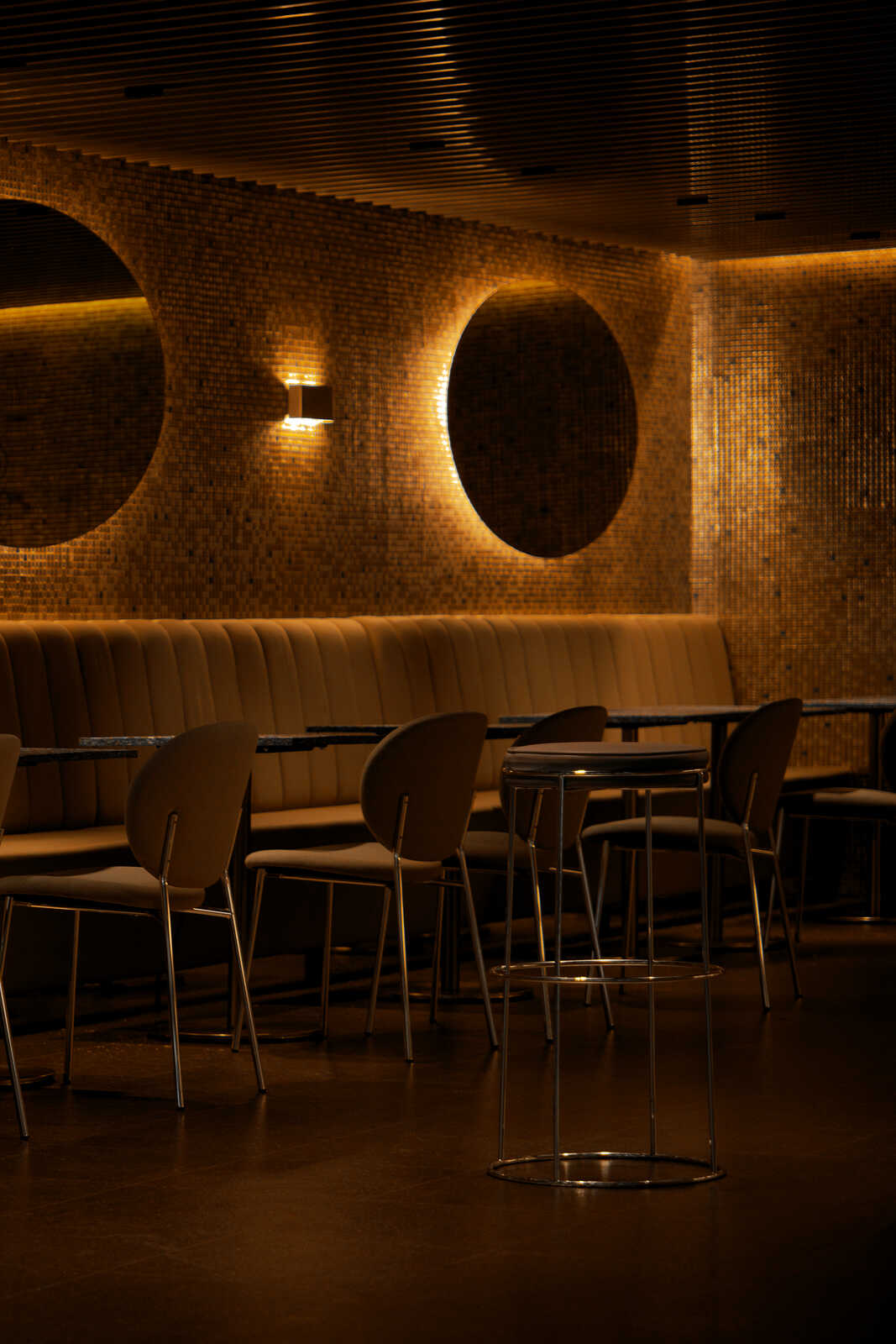 A modern interior with dark-toned seating, round tables, and chairs. There's a patterned wall with circular light fixtures. The scene is dimly lit, creating a warm atmosphere.