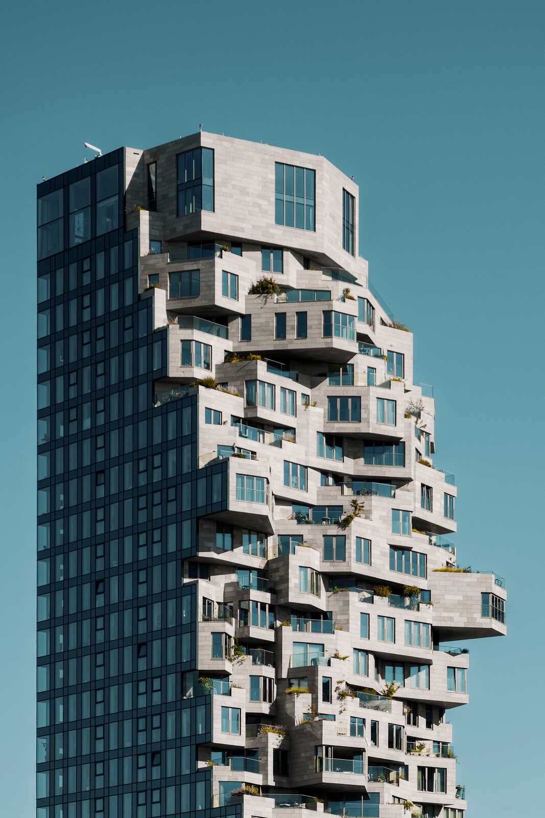 A tall, modern building with a unique, stepped design. Each level features numerous balconies with greenery. The structure is light-colored against a clear blue sky.