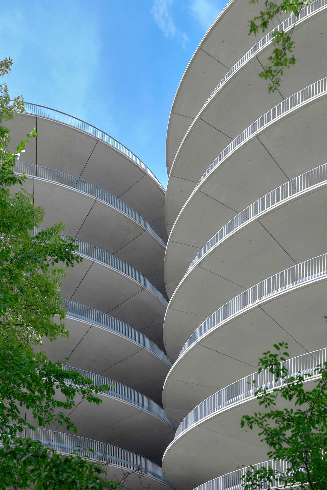 Circular concrete structure with white railings on each level. Blue sky and green foliage visible in the background. The architecture appears modern and layered.