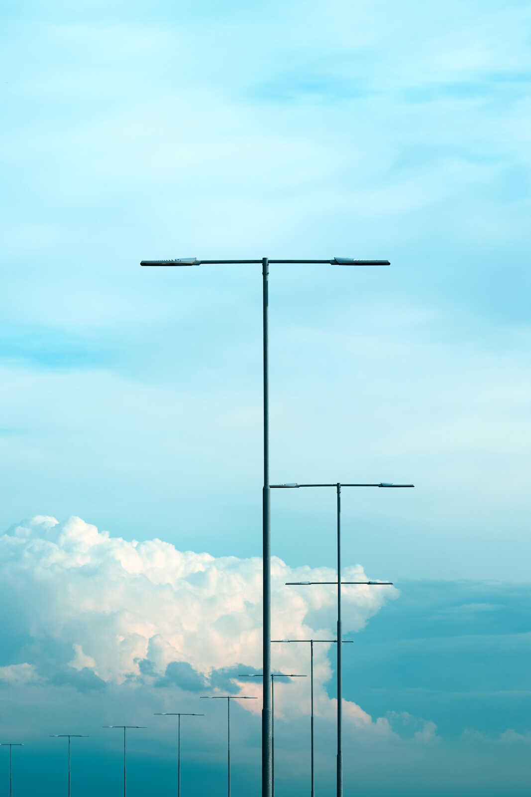 Tall, silver utility poles extend upward against a backdrop of a light blue sky with fluffy white clouds. Several poles are visible, diminishing in size towards the horizon.