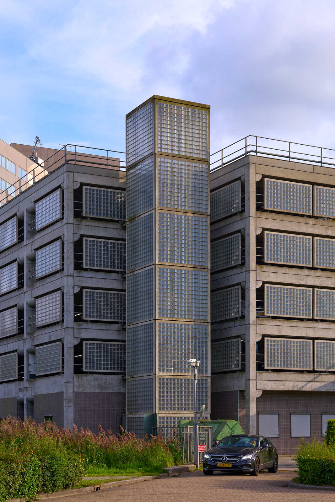 A modern gray building with a glass-block facade and a parking lot in front. A black car is parked nearby, and some green foliage grows along the base. The sky is overcast.