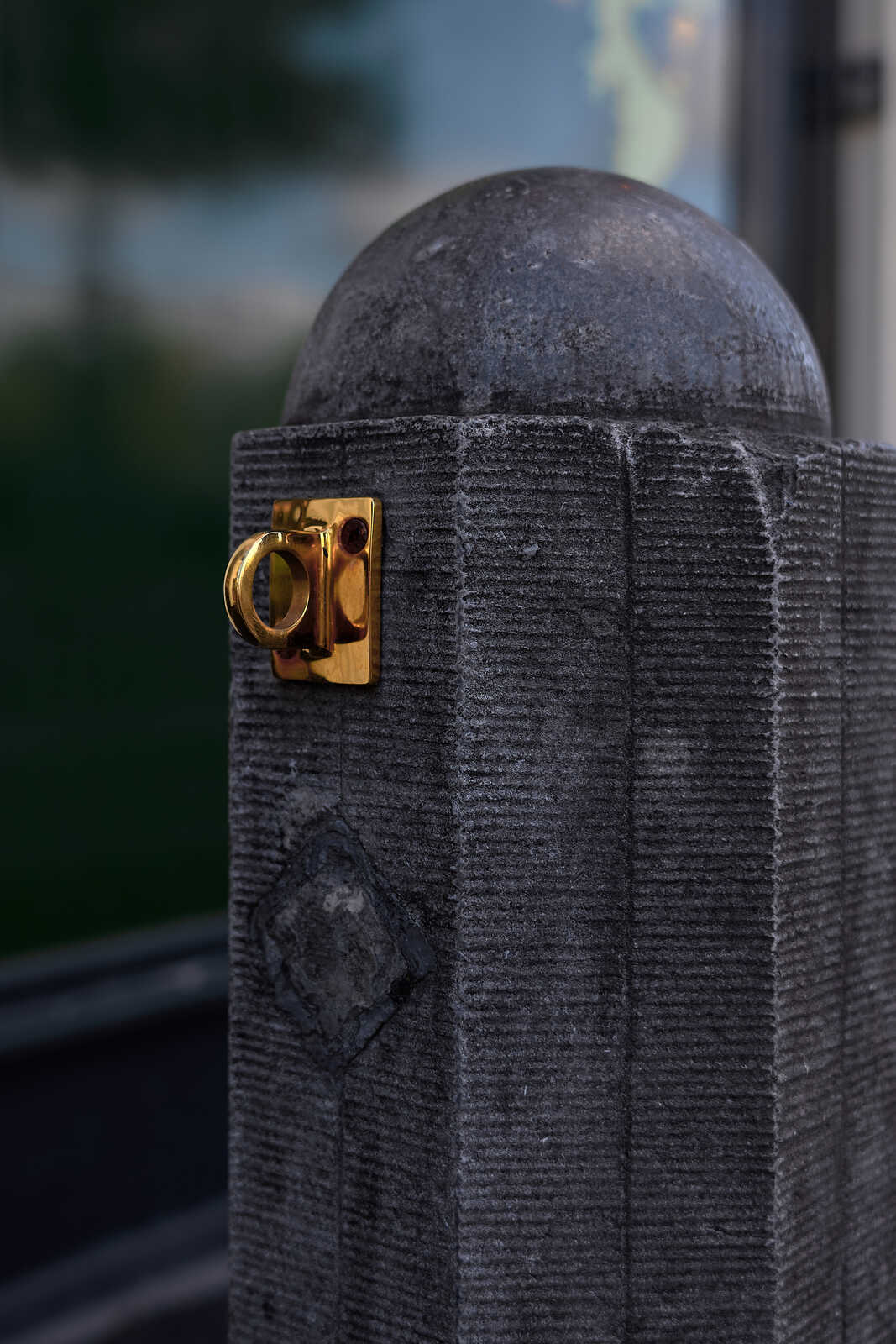 Close-up of a gray textured pillar with a brass ring attached. The ring is secured by a square plate, and the pillar has vertical grooves. The background is blurred and dark gray/black.
