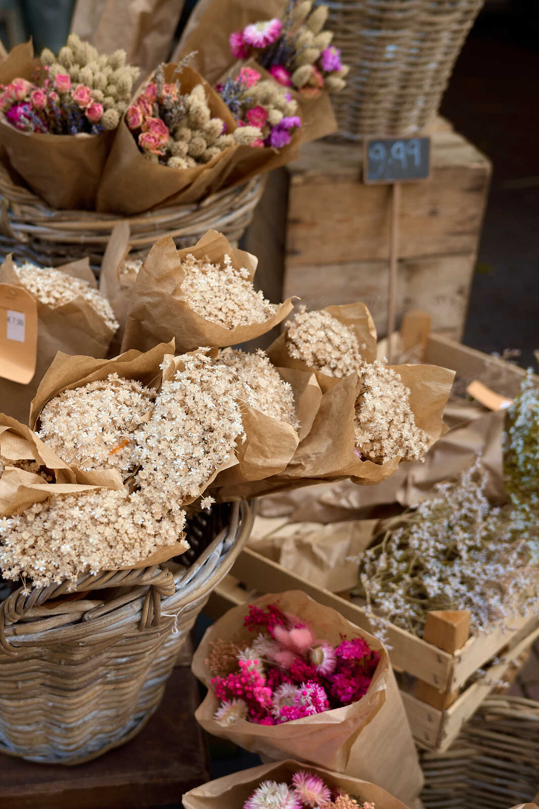 Several bouquets of dried flowers wrapped in brown paper sit on a wooden shelf. A wicker basket and a small sign are also visible.