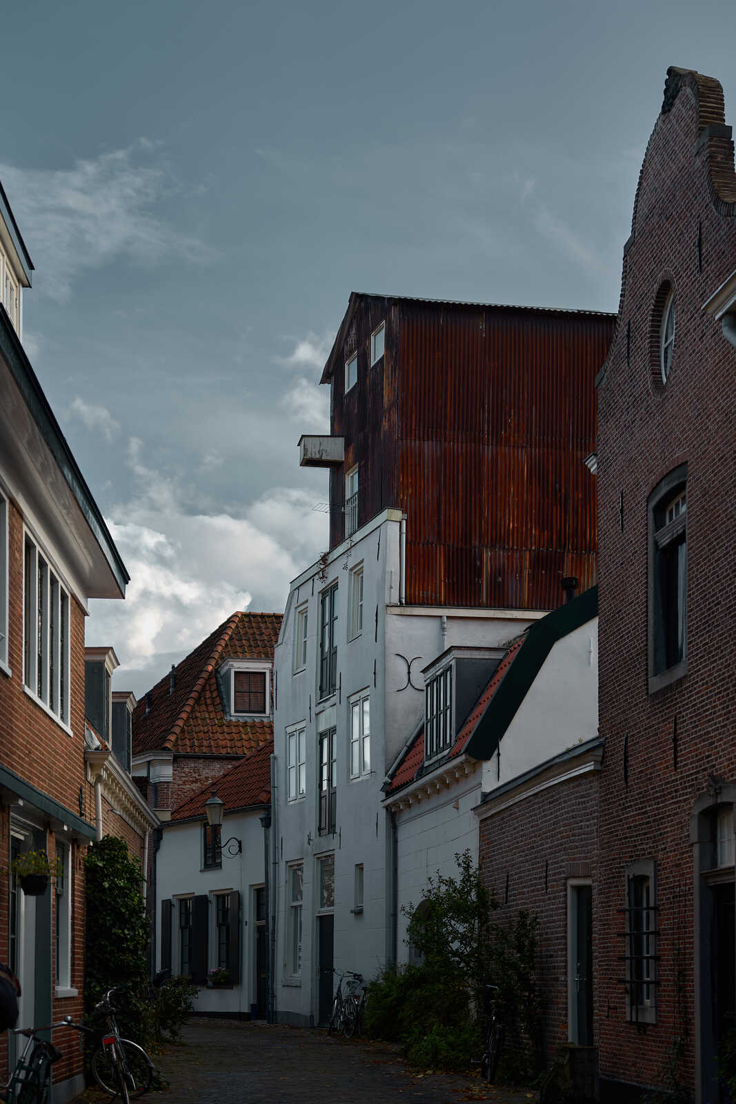A narrow street with white and red brick buildings. A tall metal structure with a corrugated roof stands at the end. Bicycles are parked along the side. Overcast sky above.