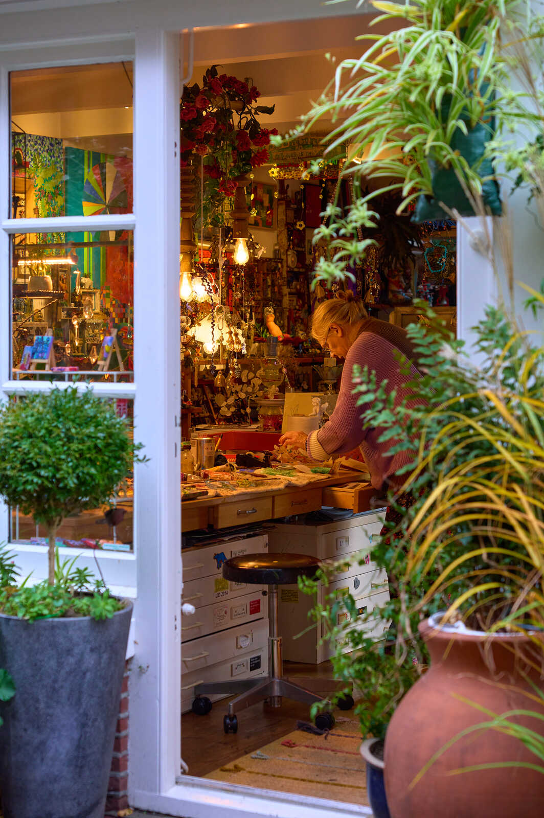 A person wearing a pink sweater stands at a cluttered table filled with colorful beads and crafting supplies. Potted plants sit on the floor and window sills. A large window provides natural light.