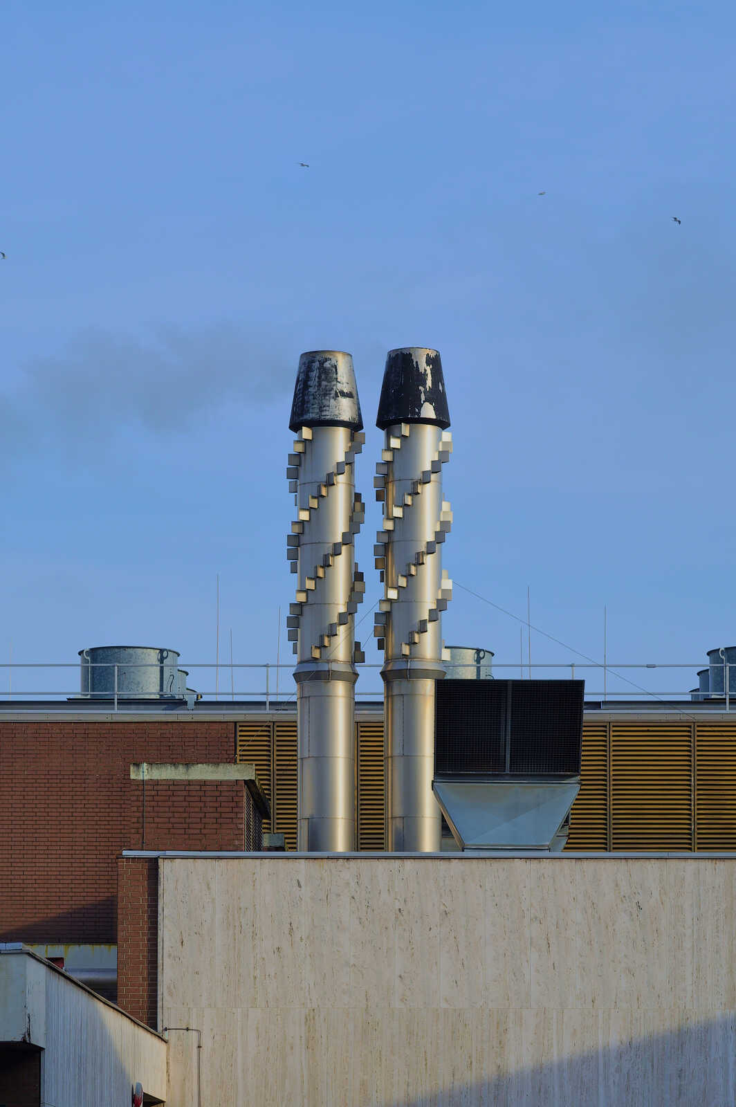Two tall, silver, spiraling vents rise above a beige and brick building. A clear blue sky fills the background. Several vents and rooftop equipment are visible below.