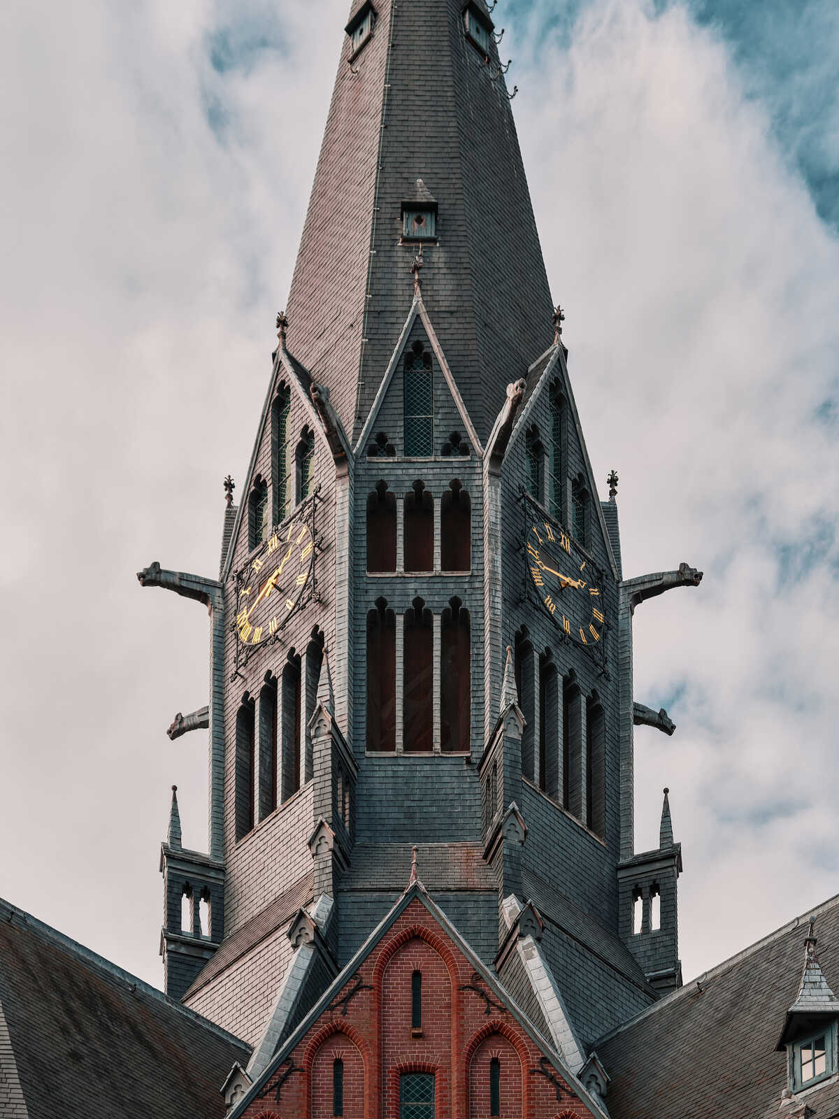 Tall gray stone tower with a clock face. Gothic arched windows and ornate details. A cloudy sky fills the background. Red brick base visible below the stone facade.