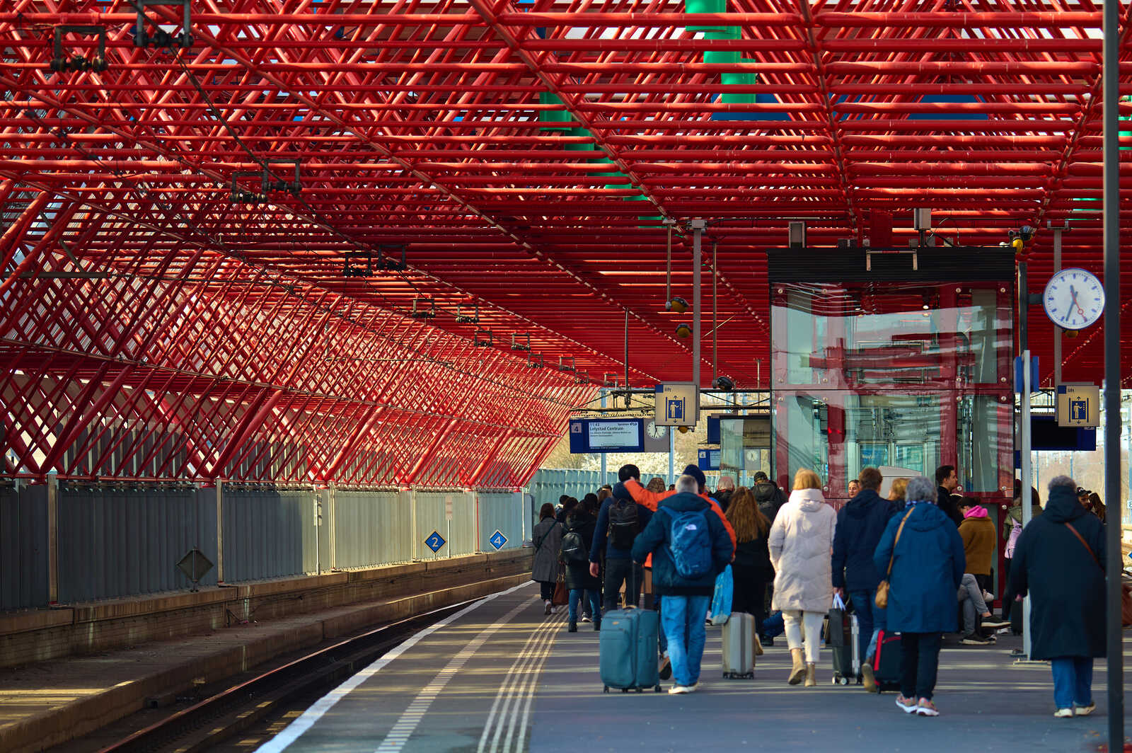 People are walking along a platform with a red, grid-like roof. Theres a clock and signage on the wall. Suitcases are visible among the crowd. A train track is present.