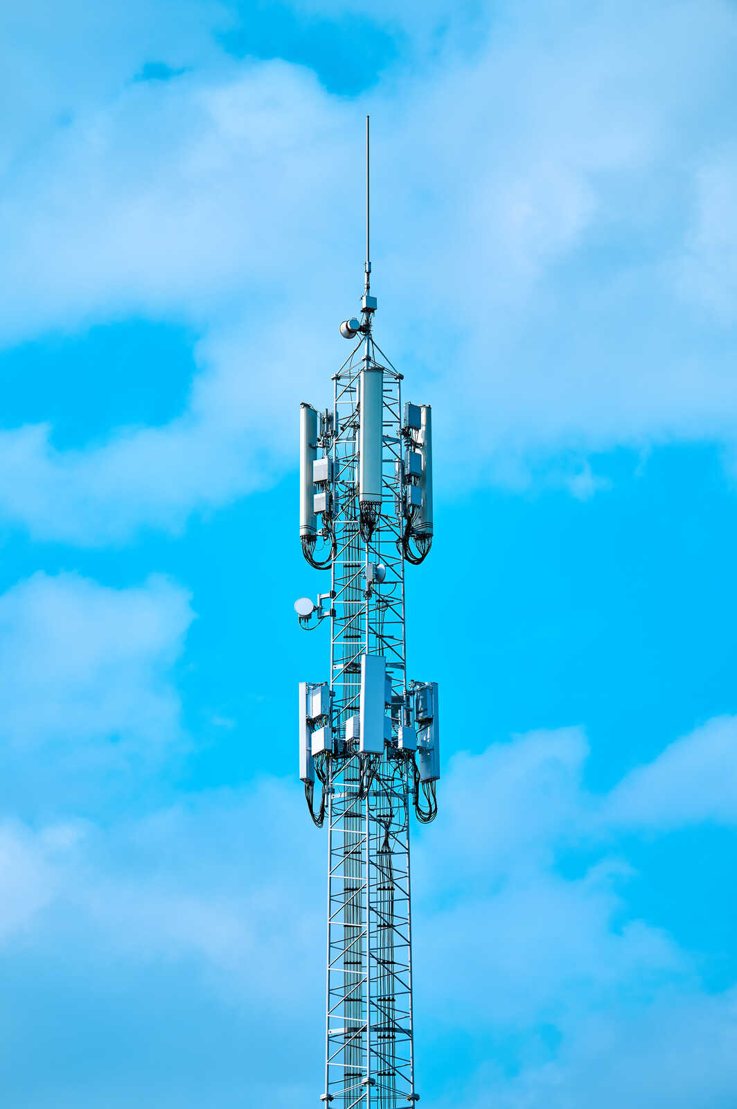 A tall, gray communication tower with multiple antennas and dishes extends against a bright blue sky with scattered white clouds. It has a central mast with cross bracing.