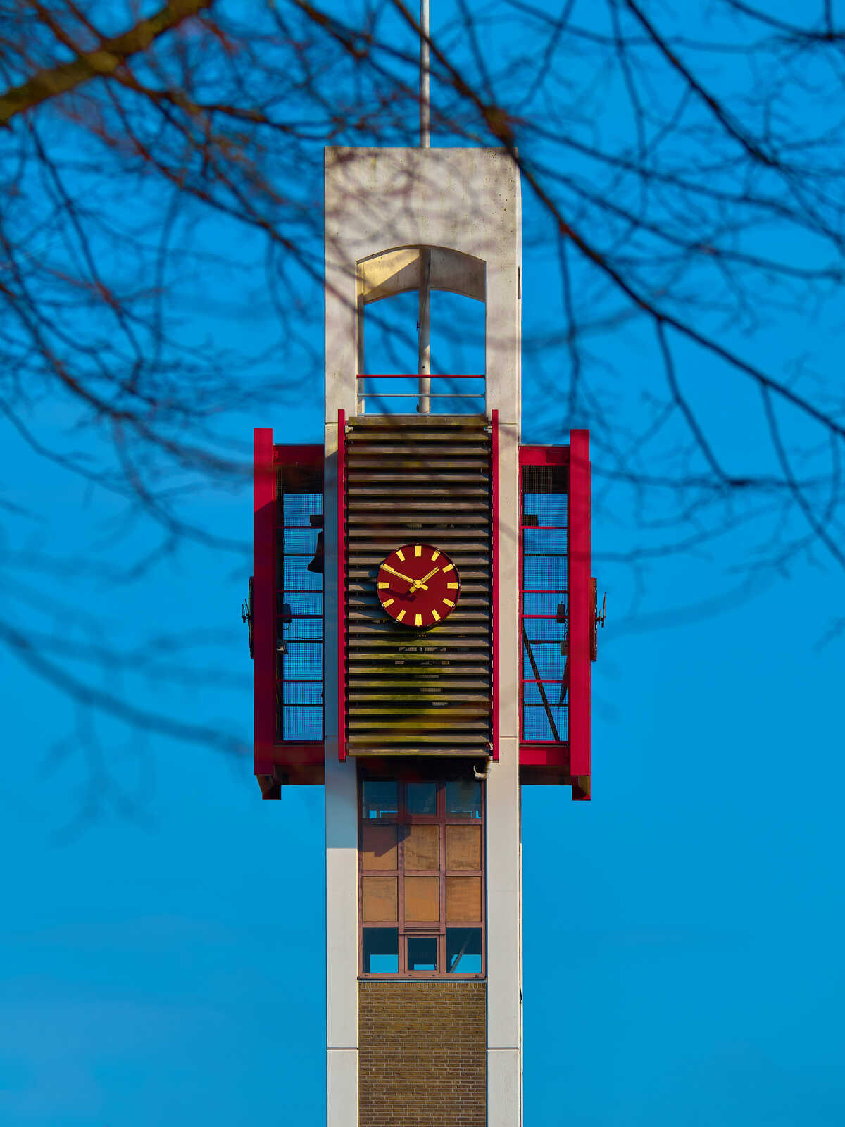 Tall, gray tower with a clock face. Red fire escape railing. Brick base. Blue sky background. Minimalist architecture. Vertical composition. Tree branches visible at the top.