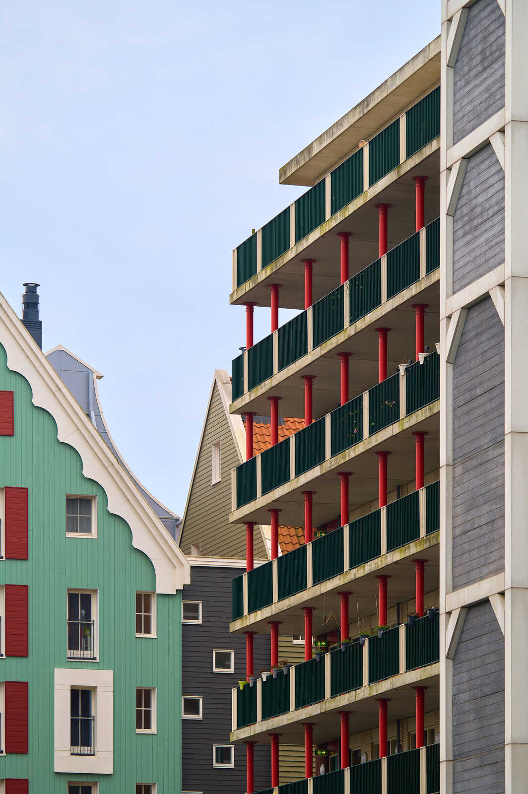 Gray and green buildings are shown. The building on the right has a balcony with red pillars and green railings. The building on the left has windows and shutters.
