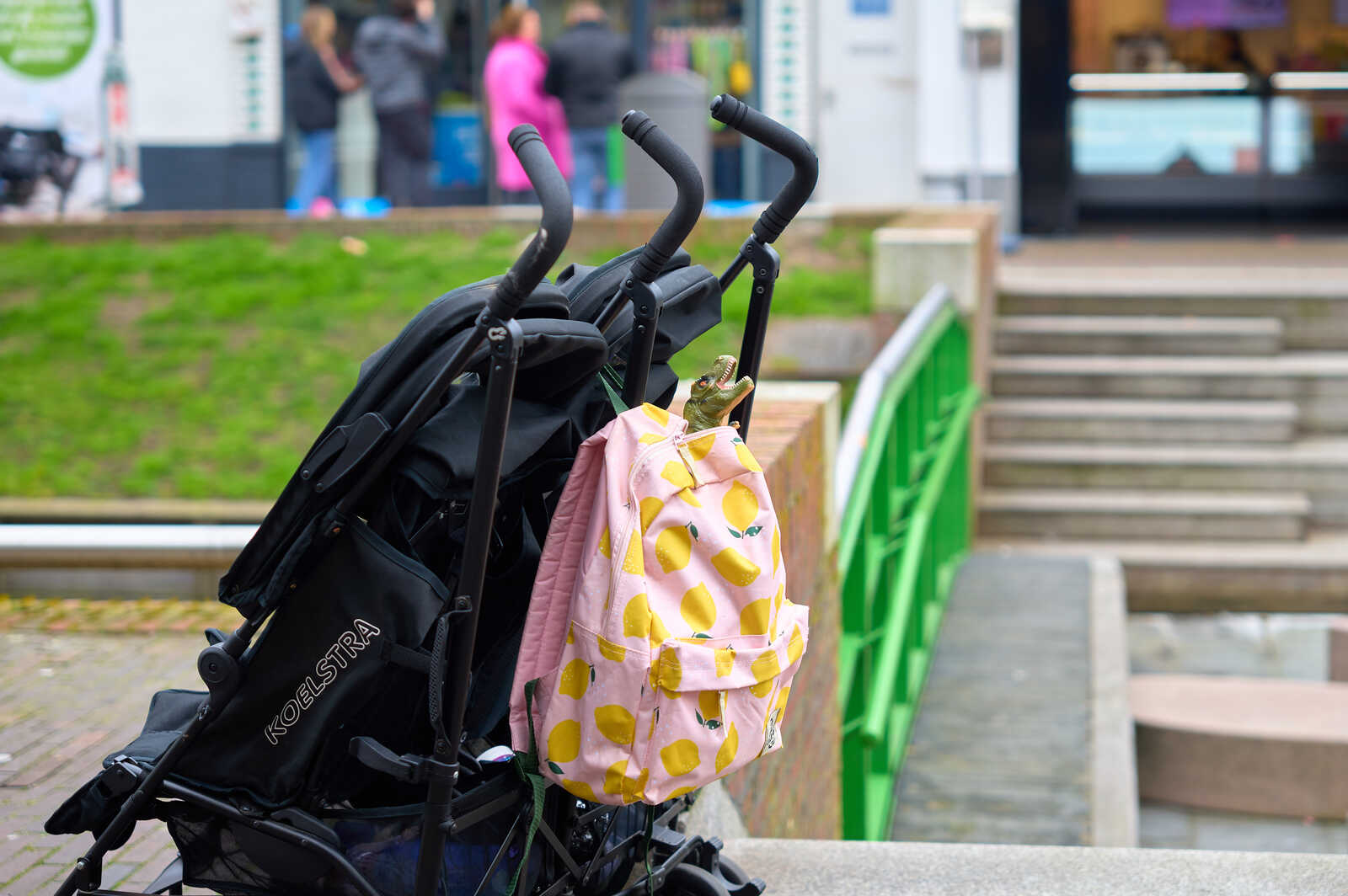A black stroller with a yellow backpack hanging from the handles. The backpack features a lemon pattern. There are gray steps and a green railing in the background.