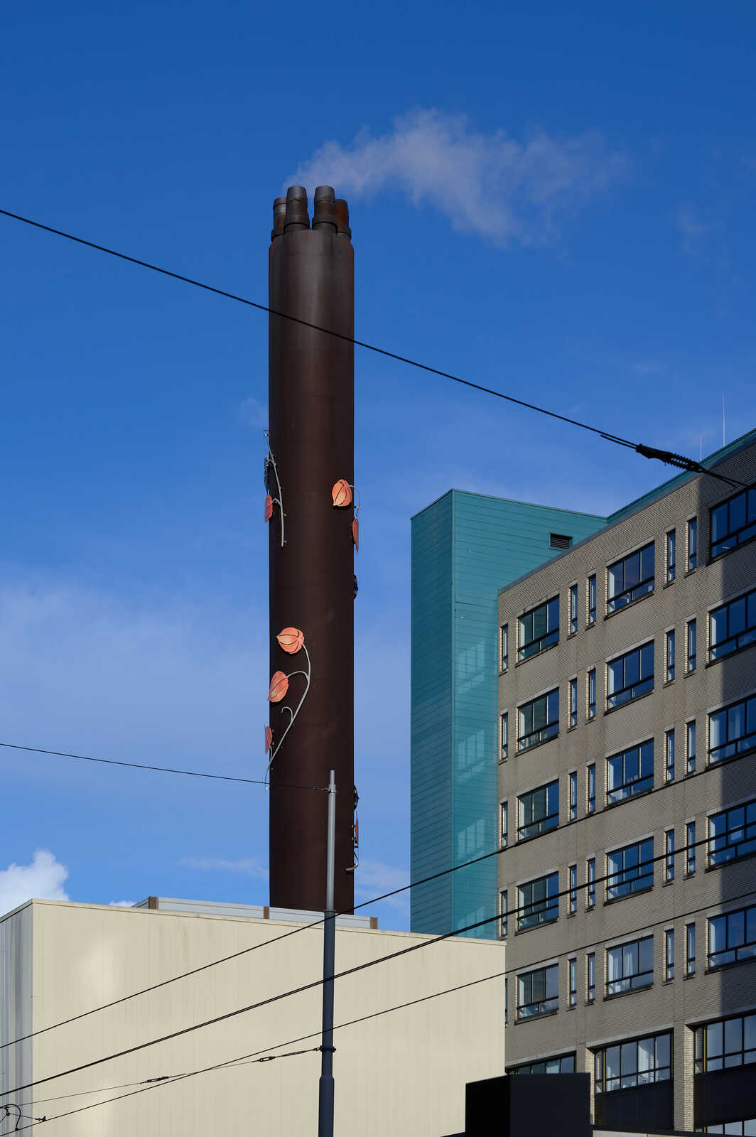 A tall, brown chimney with decorative copper-colored leaves ascends against a blue sky and a modern, multi-story building. A power line runs across the top of the frame.