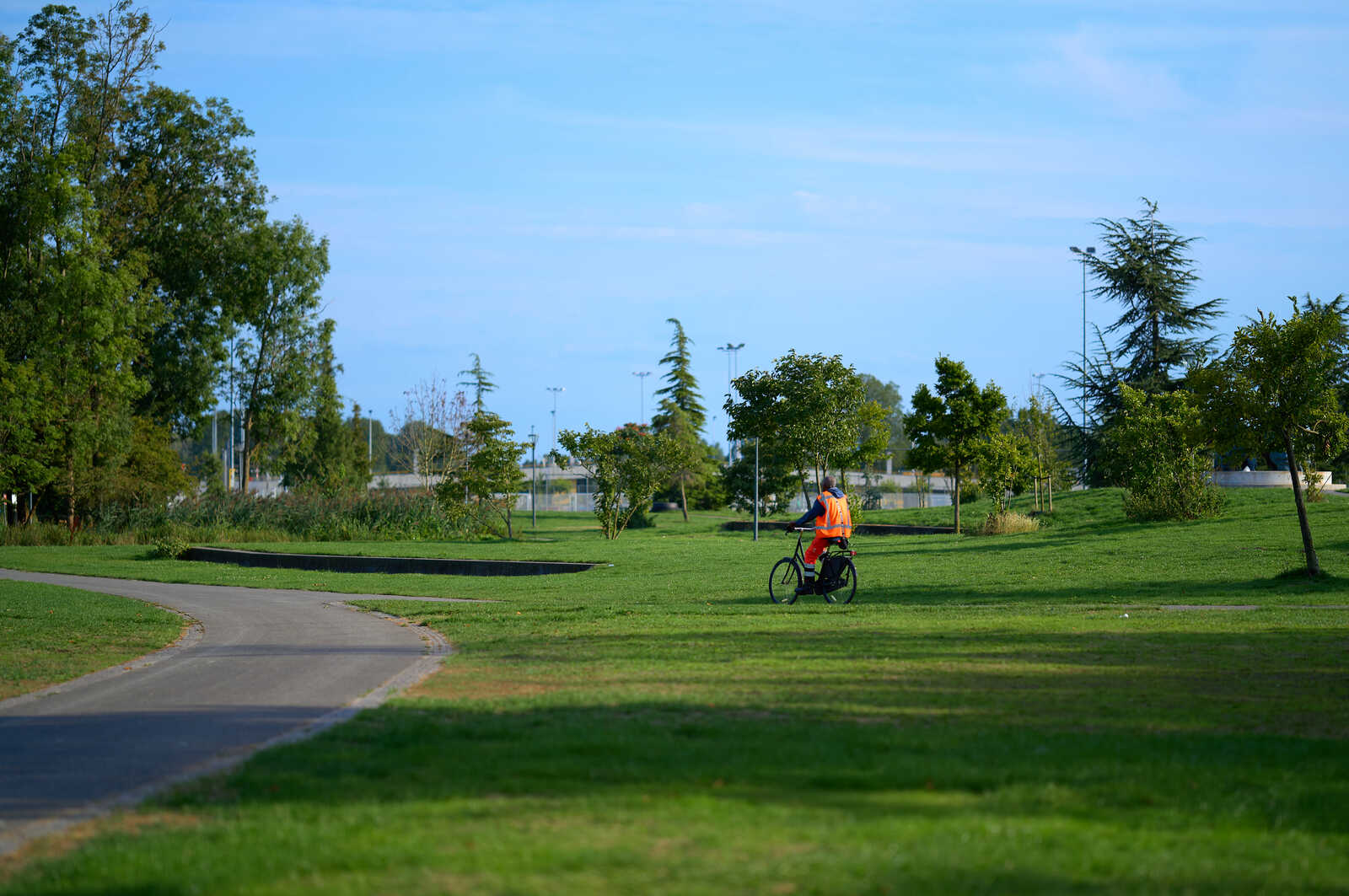A person in an orange vest rides a bicycle on a paved path. Lush green grass and trees fill the background under a clear blue sky. A pond is visible in the distance.