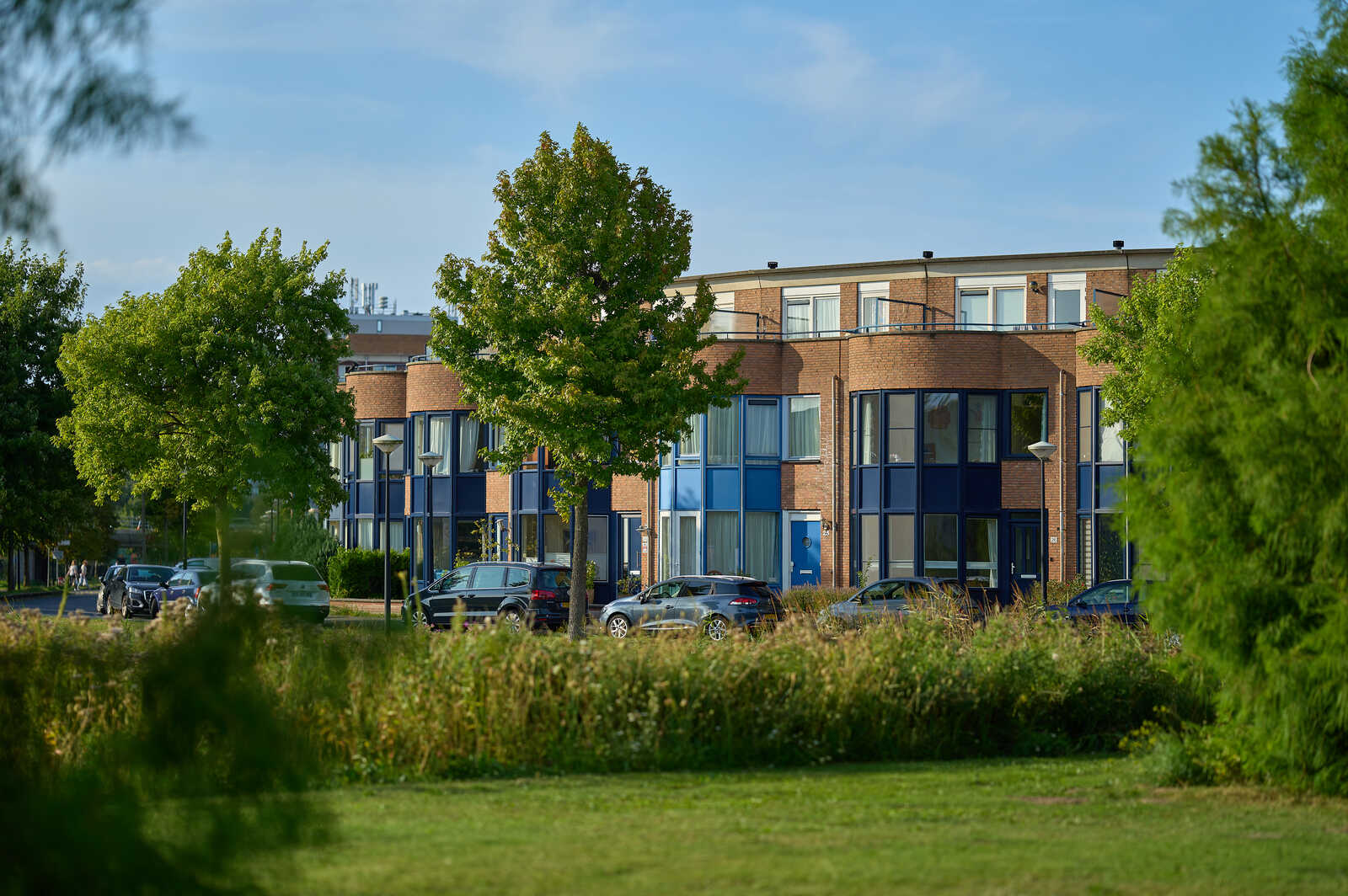 Red brick apartment building with blue window frames and balconies. Grass and greenery in the foreground. Several cars parked in front of the building. Blue sky overhead.