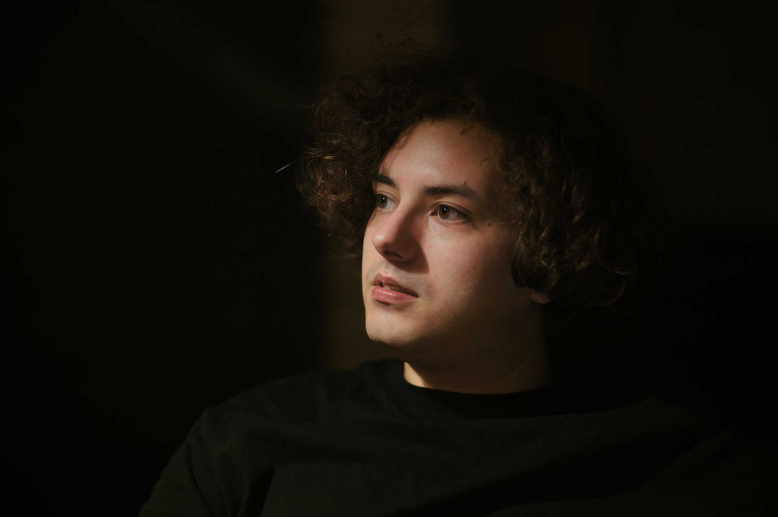 A young man with curly dark hair and a dark shirt looks off-camera. He has fair skin and is lit from a light source to his right. His expression is neutral. Black background.