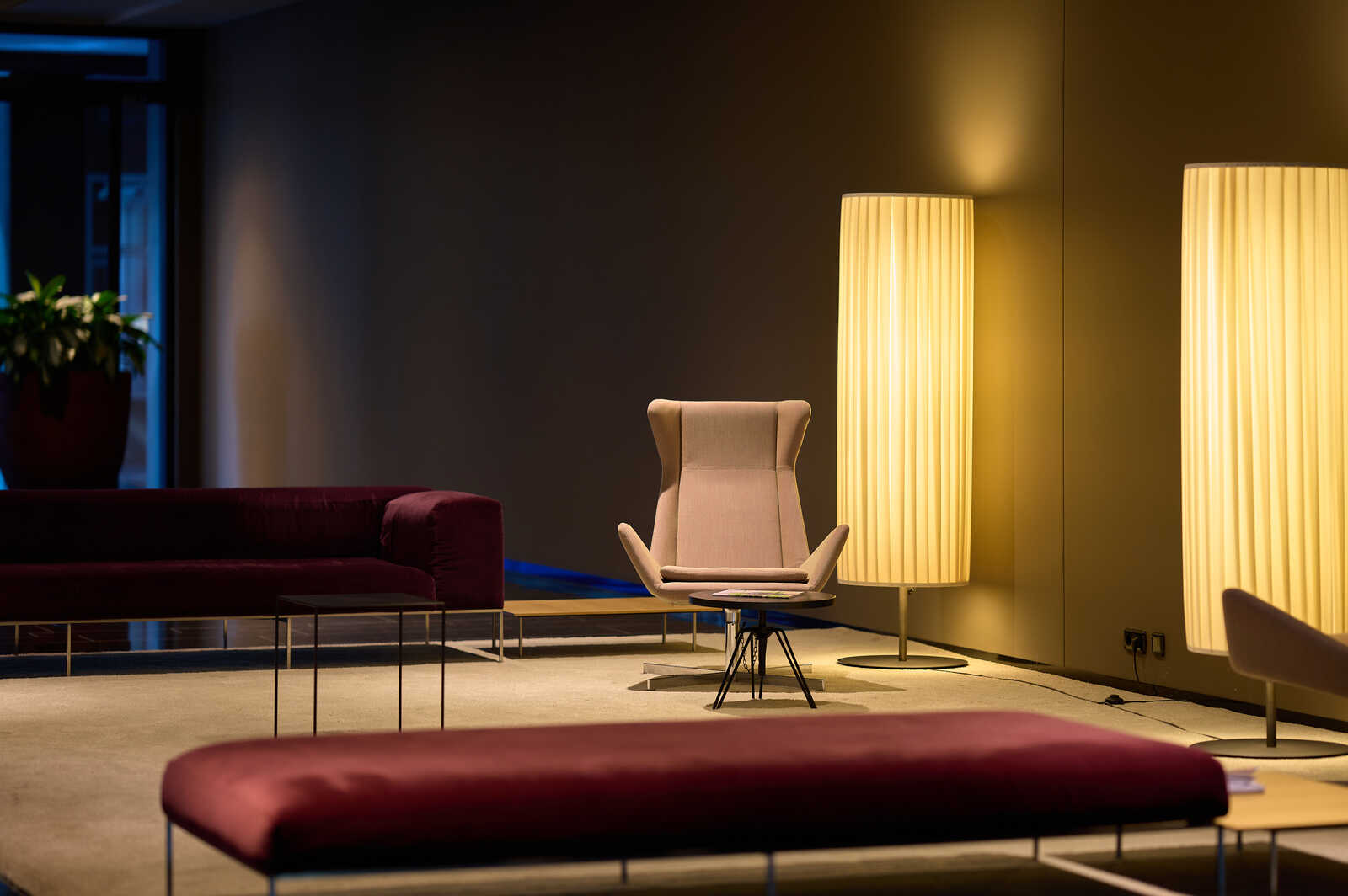 A room with a red couch, ottoman, chair, and tall floor lamp. A potted plant sits in the background near a window. The floor is carpeted in a neutral tone.