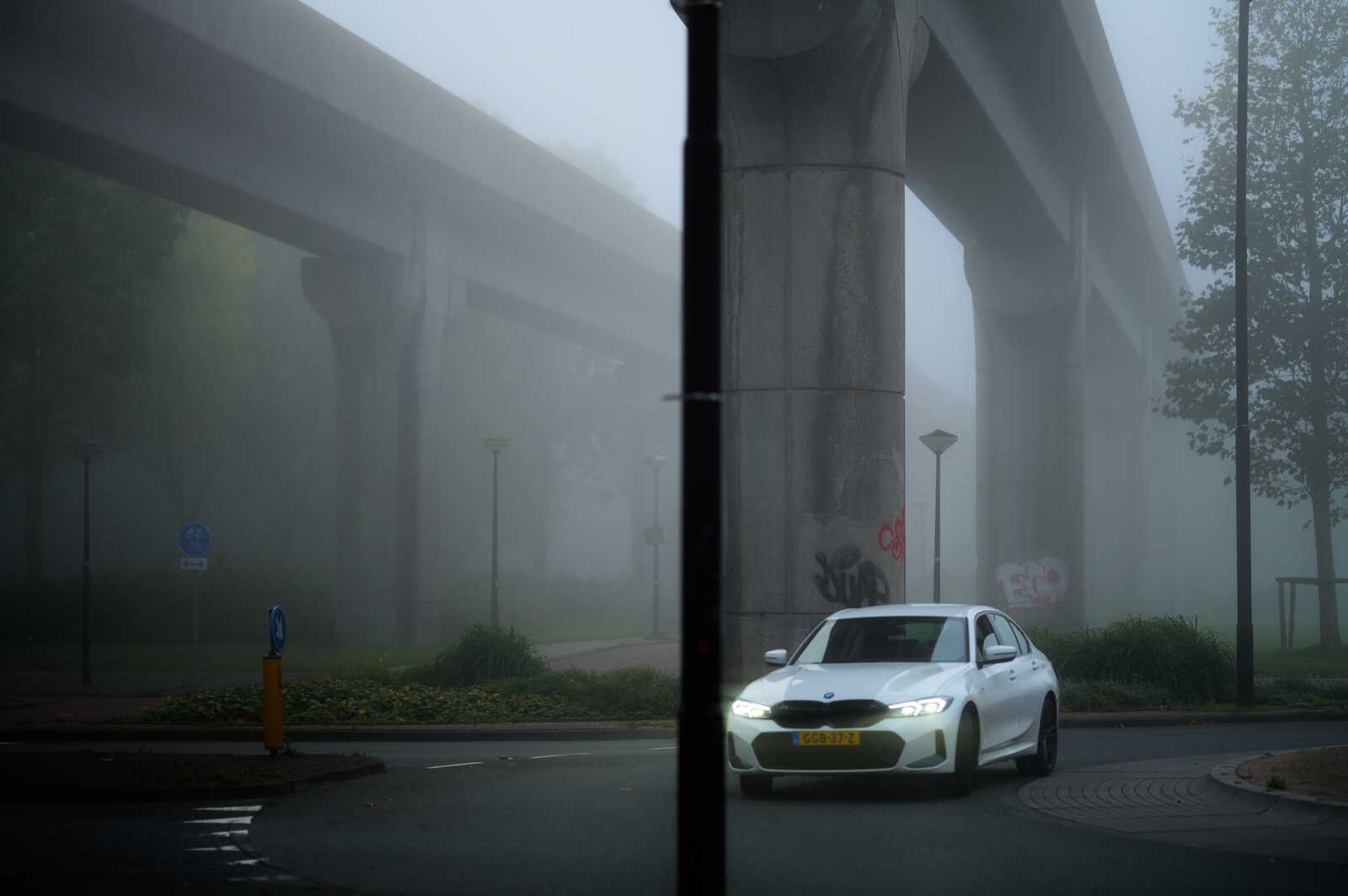 White BMW sedan on wet pavement. A bridge structure and foliage are visible in heavy fog. A traffic sign and a yellow pole are on the left. Graffiti is on the bridge wall. Road markings are present.