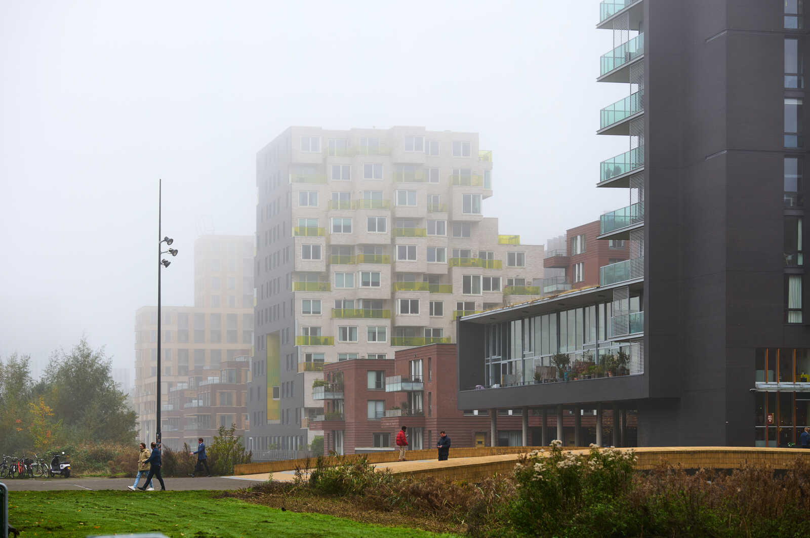 A modern apartment building with balconies is visible through fog. Several people are walking on a path near a grassy area and a bike rack.