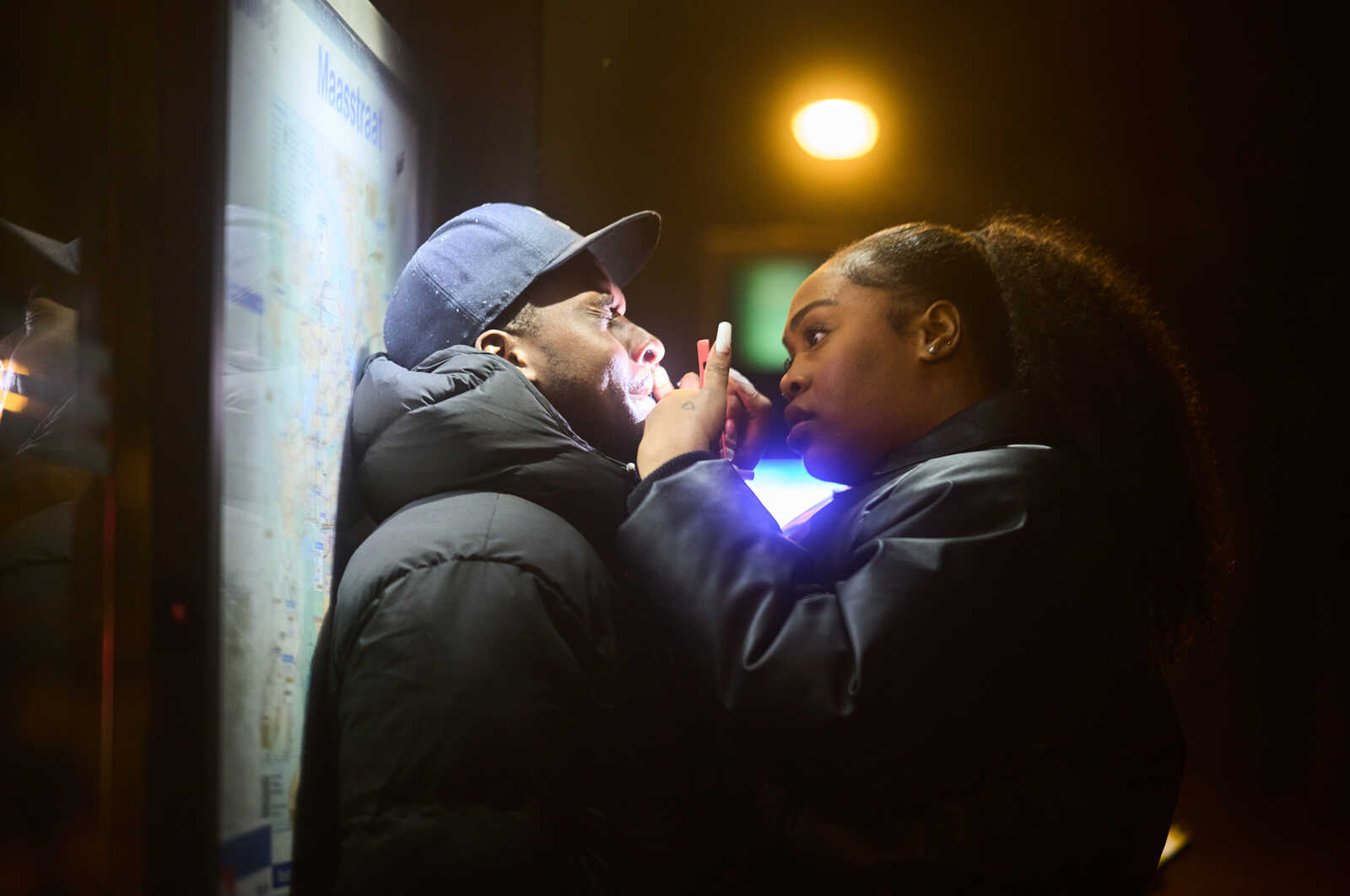 Two people stand close, facing a brightly lit screen. One wears a black puffer jacket and a baseball cap, while the other has dark hair and a black coat. They seem to be looking at the screen together.