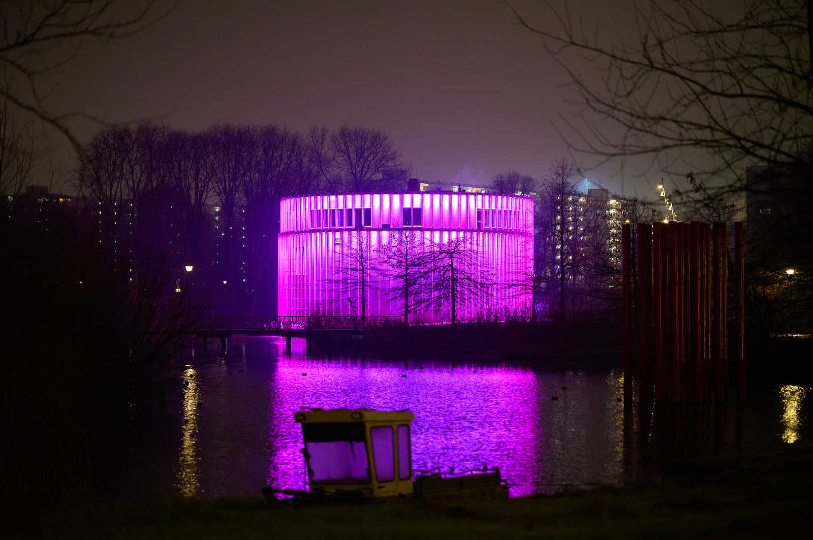 A round building illuminated in purple light reflects on still water. A boat sits on the bank, with a red structure and dark trees visible in the background.